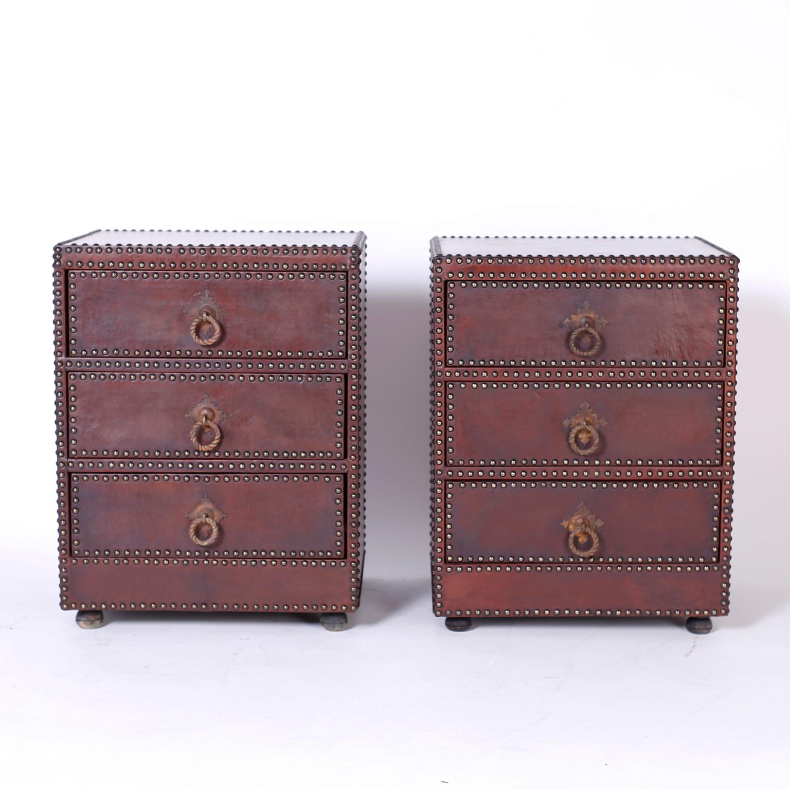 Handsome pair of three drawer chests or stands, entirely clad in brown leather highlighted with tacks, handwrought metal hardware and bun feet.