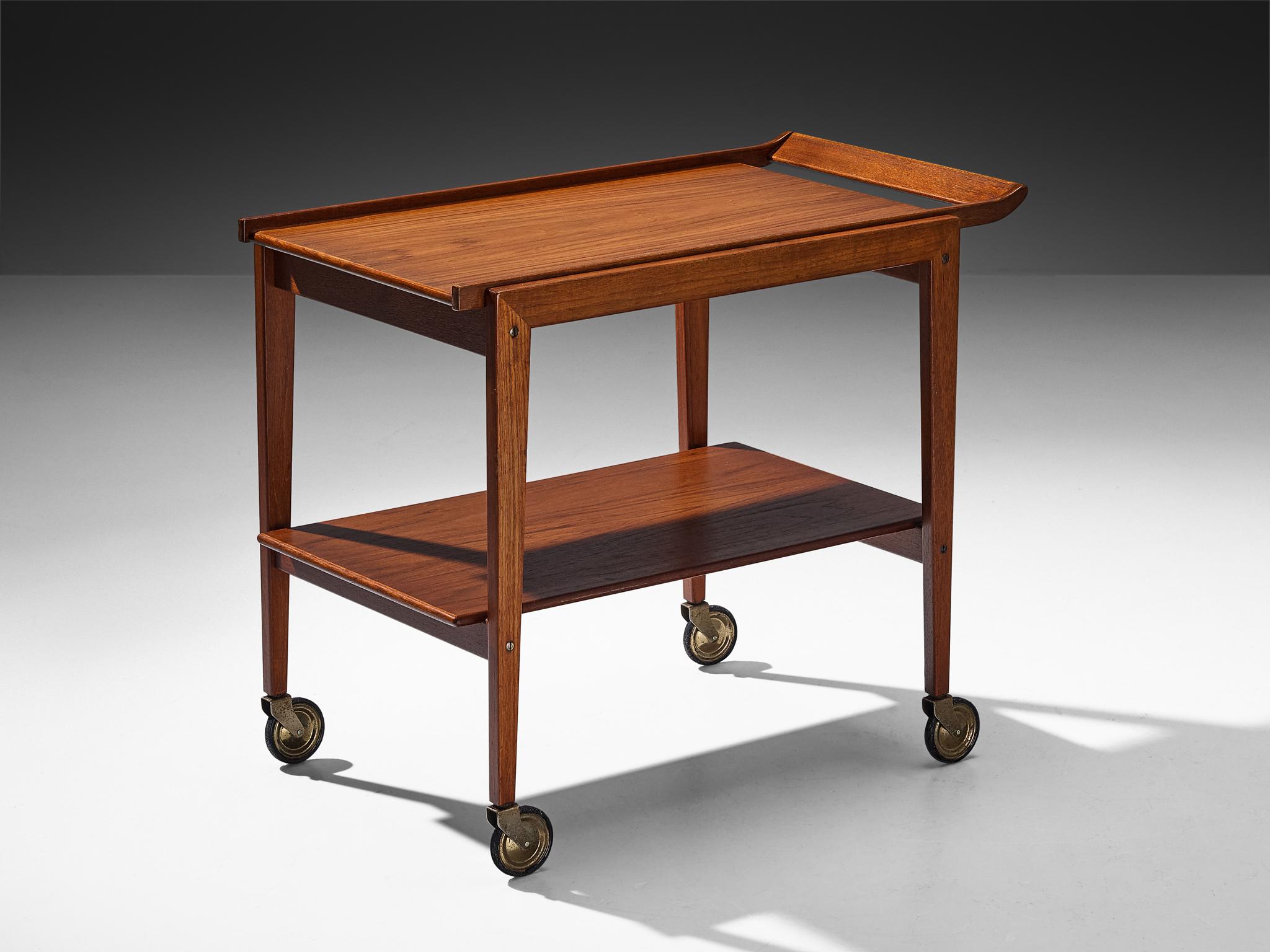 Trolley or bar cart, teak, brass, Europe, 1960s.

This elegant bar cart is crafted from teak, featuring a sturdy wooden frame and two spacious shelves. Set on four wheels, it doubles as a functional trolley, offering both style and mobility. The