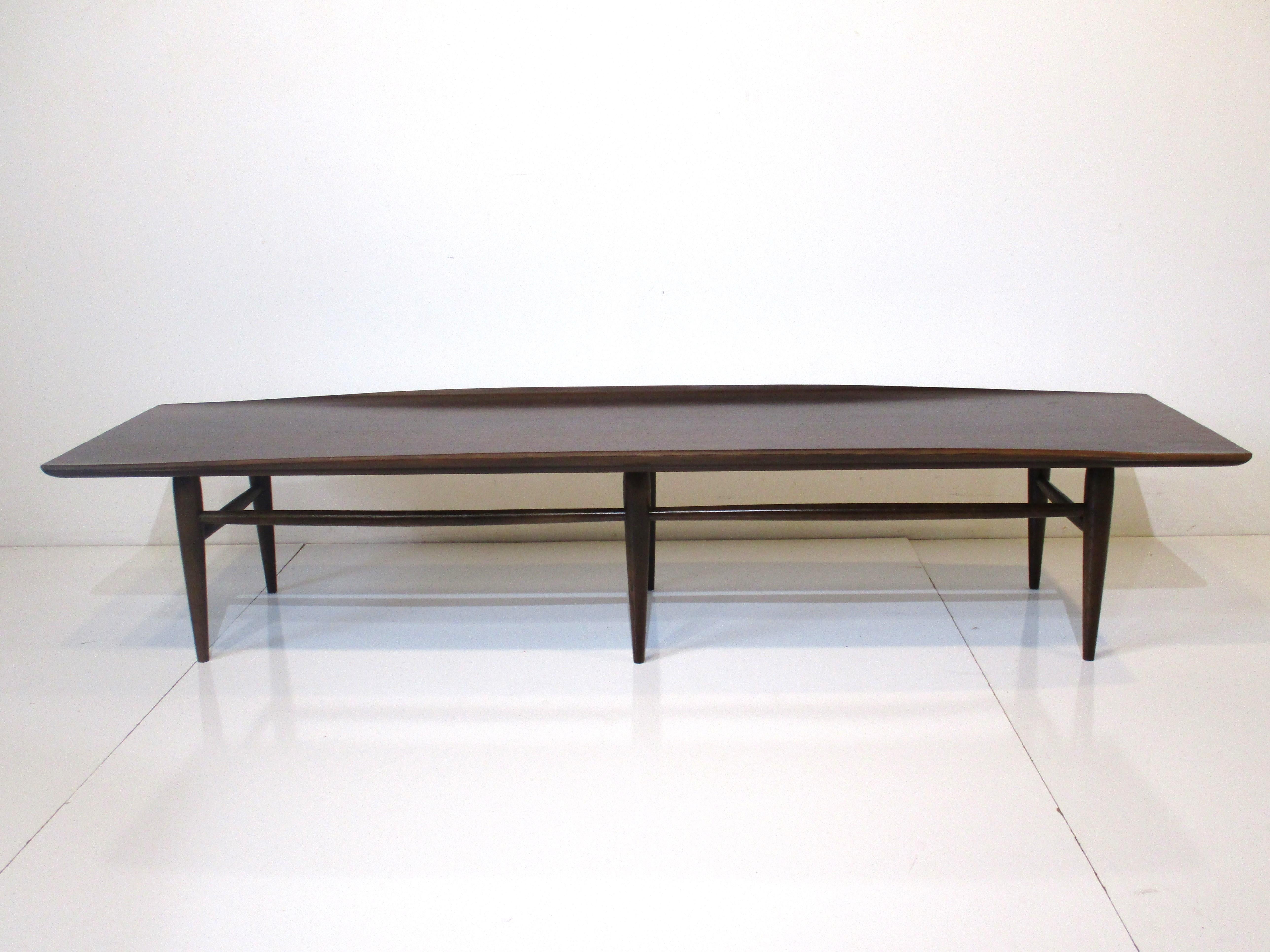 A long medium dark ebony toned wooden coffee table with up turned edges to both sides sitting on conical legs and stretches' to each one. Manufactured by the Drexel Furniture company, well made and the perfect piece for that Mid Century room.