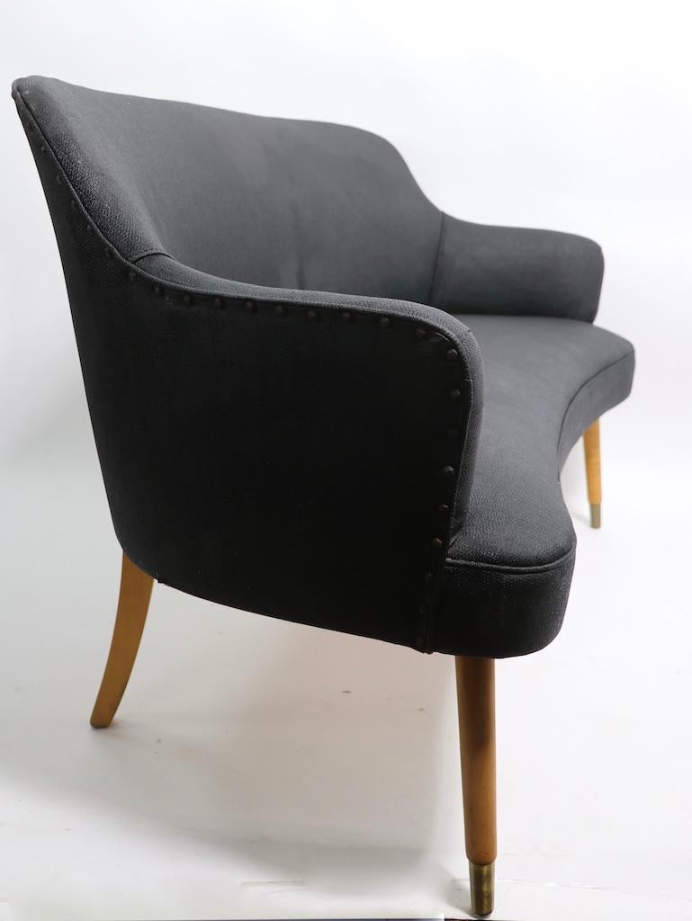 Chic and stylish organic kidney shaped loveseat attributed to chair master. The seat and back are upholstered in very dark gray textured vinyl, the taped pole legs are of solid wood, with brass capped front feet. The seat has a minor flaw (tear),