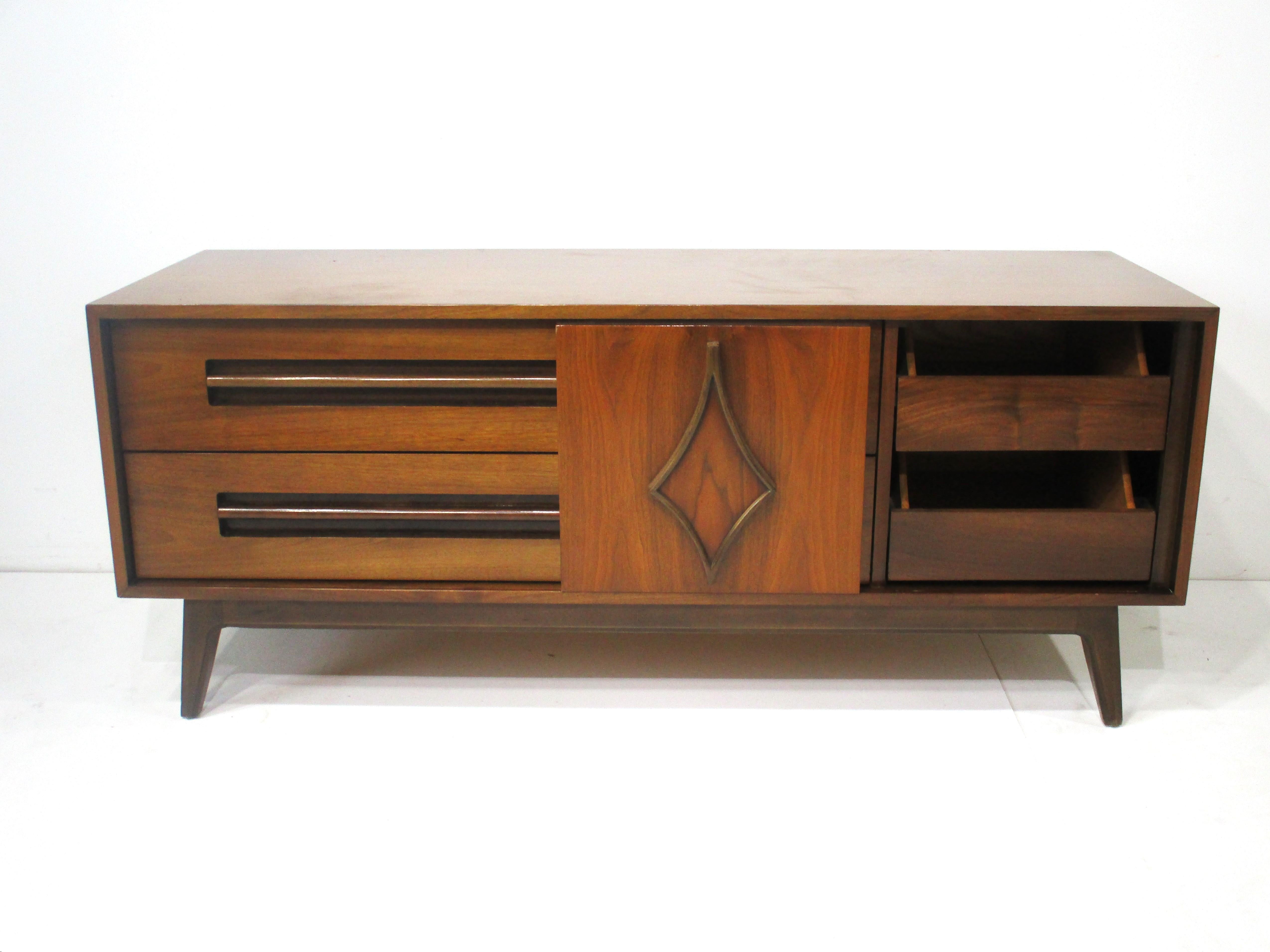 A low profile walnut credenza with a sculptural sliding door covering two smaller drawers and two longer drawers to the other side . The long pulls have a cut out finger pulls to the bottom of each handles . With a wood trimmed design to the front