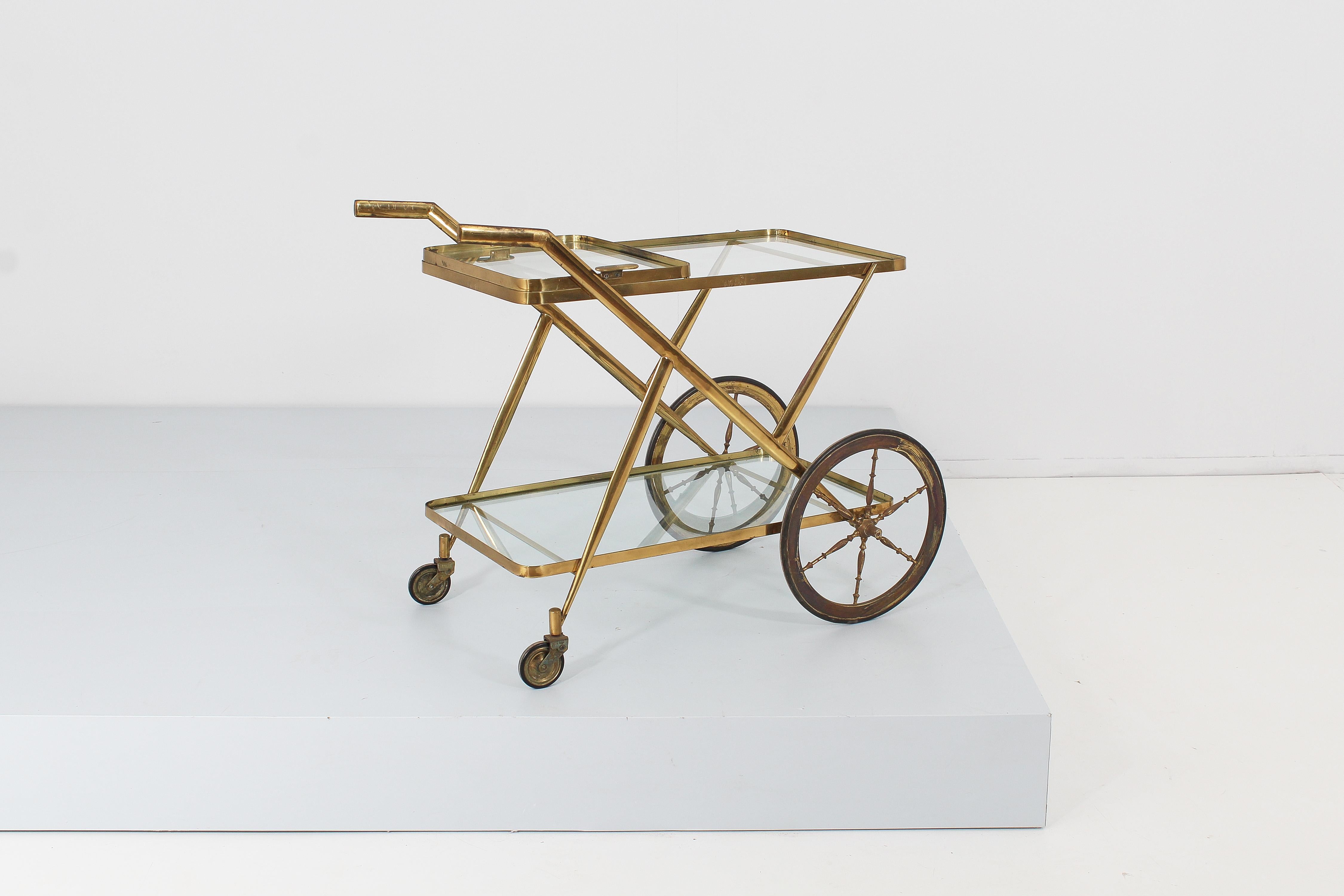 Elegant bar cart with geometric lines in solid brass with two glass shelves and two pairs of wheels with different sizes.
The small glass tray with brass profile and handles can be removed from the perimeter of the upper shelf. Attributed to Luigi