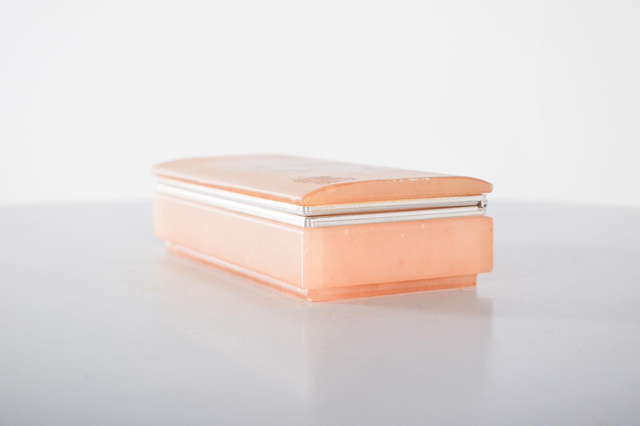 Joyero. Esta caja está hecha de un material translúcido de color melocotón suave. El diseño rectangular presenta bordes redondeados y un acabado pulido para darle un toque sofisticado. La tapa abatible está unida por bisagras de metal plateado, lo