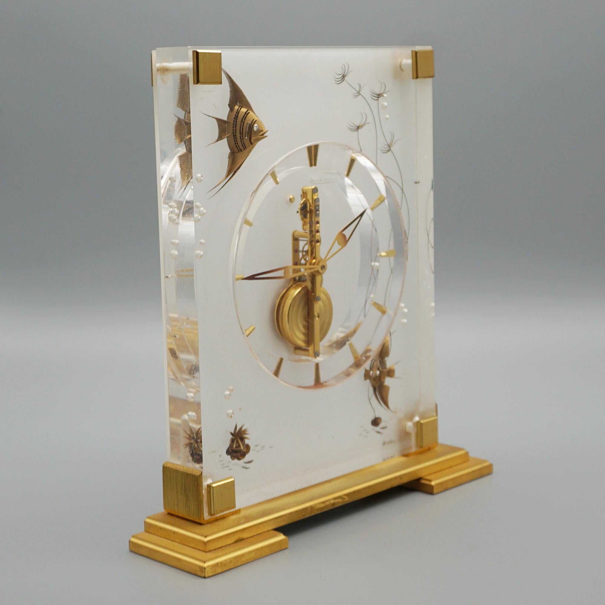 A mid-century 'Marina' mantel clock by Jaeger LeCoultre. Lucite case with inset brass decoration featuring fish amongst the reeds . Set over a gilded Brass stepped base. Eight day movement by Jaeger-LeCoultre with baton numerals and original gilded