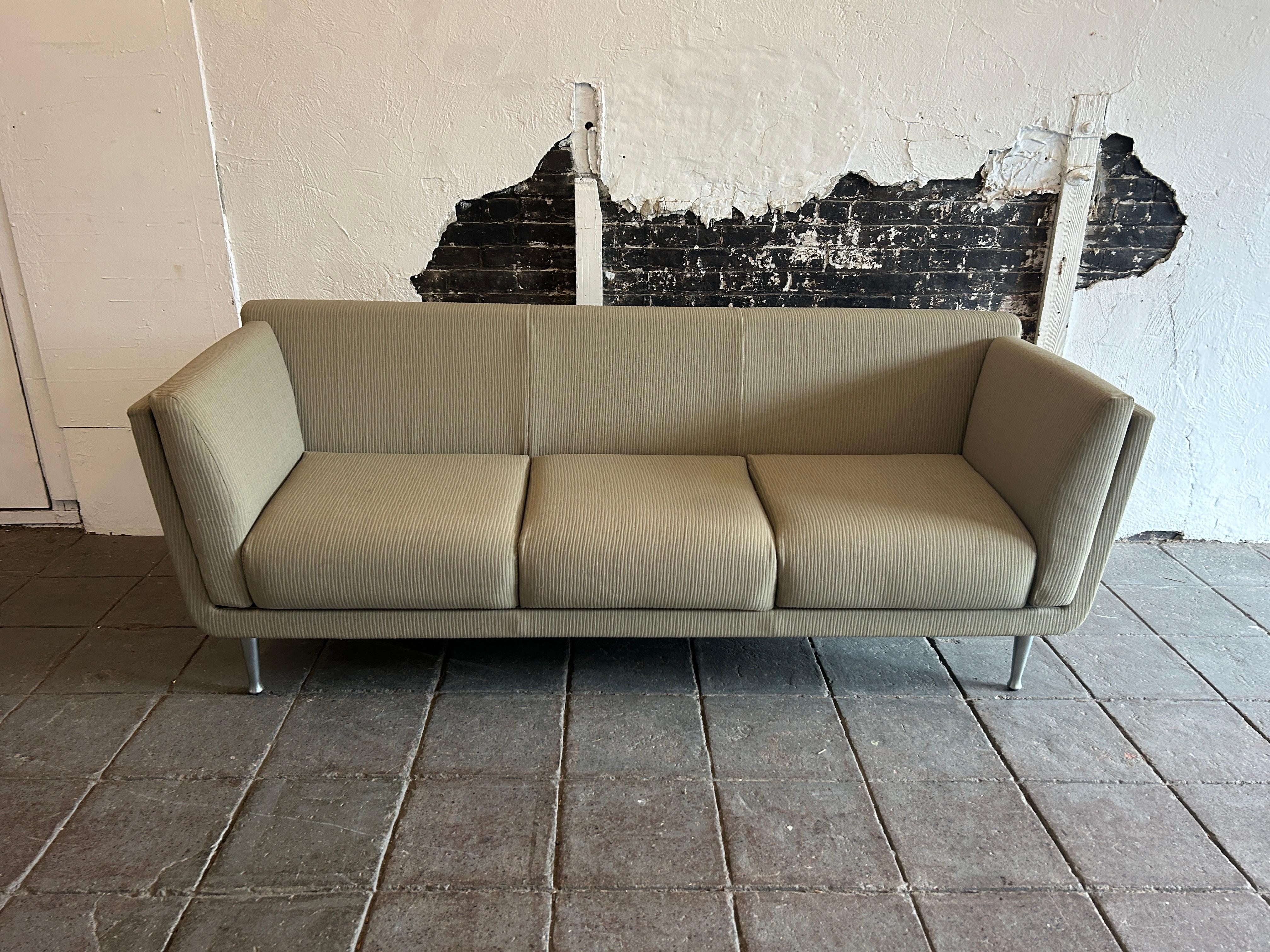 Ce canapé est recouvert d'un revêtement vert sauge/tan avec une texture verticale et une coque en bois plus clair qui repose sur 4 pieds en aluminium massif. Ce 