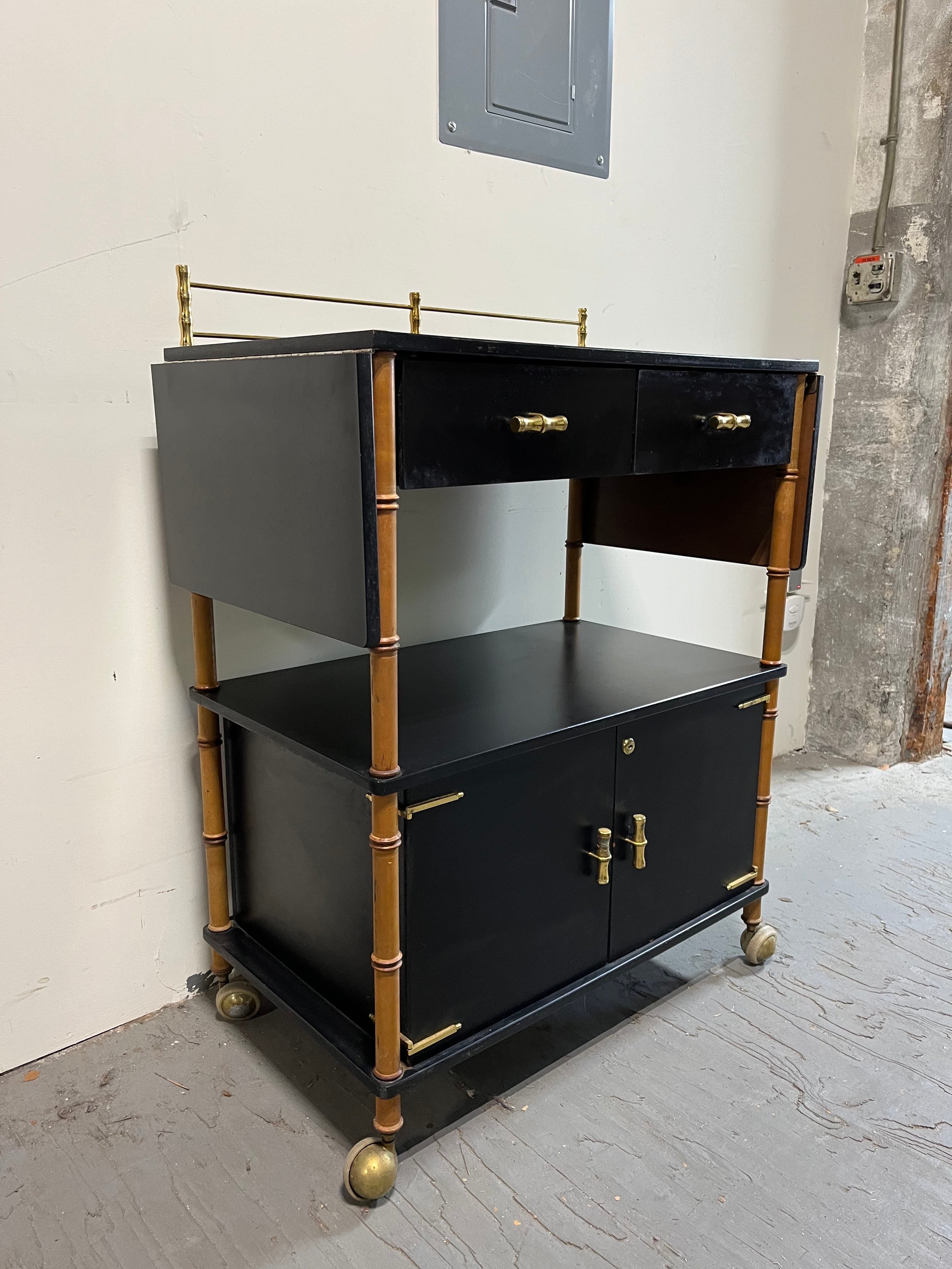 Exceptional example of high style from Maxwell Phillip, NY, NY.  Drop leaf bar cart or server. Black laminate body surrounded with epitome of classic stylized heavy brass bamboo knobs, back gallery, and hinged fasteners. Faux Bamboo legs cap off the