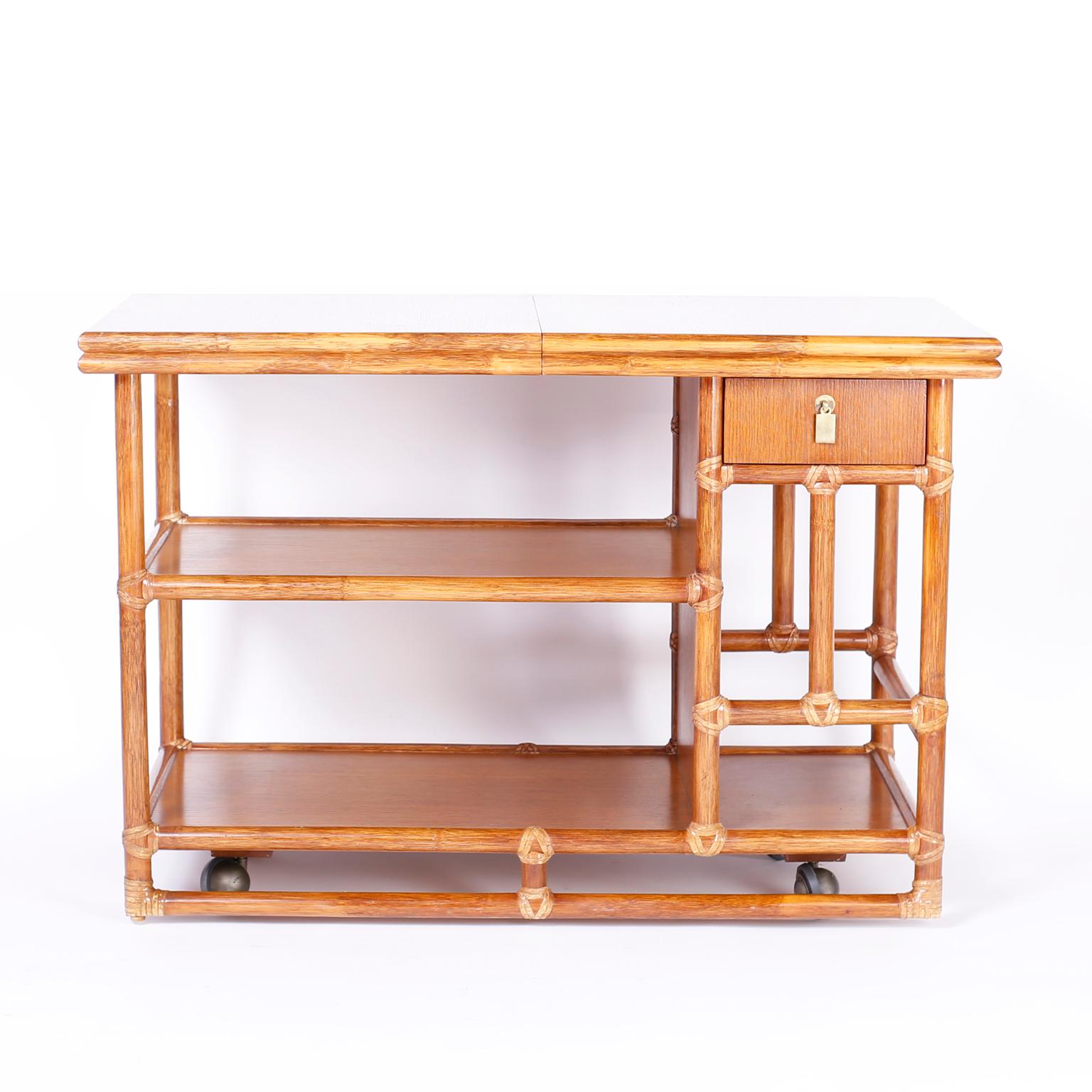 Dynamic faux bamboo bar cart with well grained oak panels, reed wrapped corners, finished all around, recessed casters and slide out service space. Signed McGuire in the drawer.