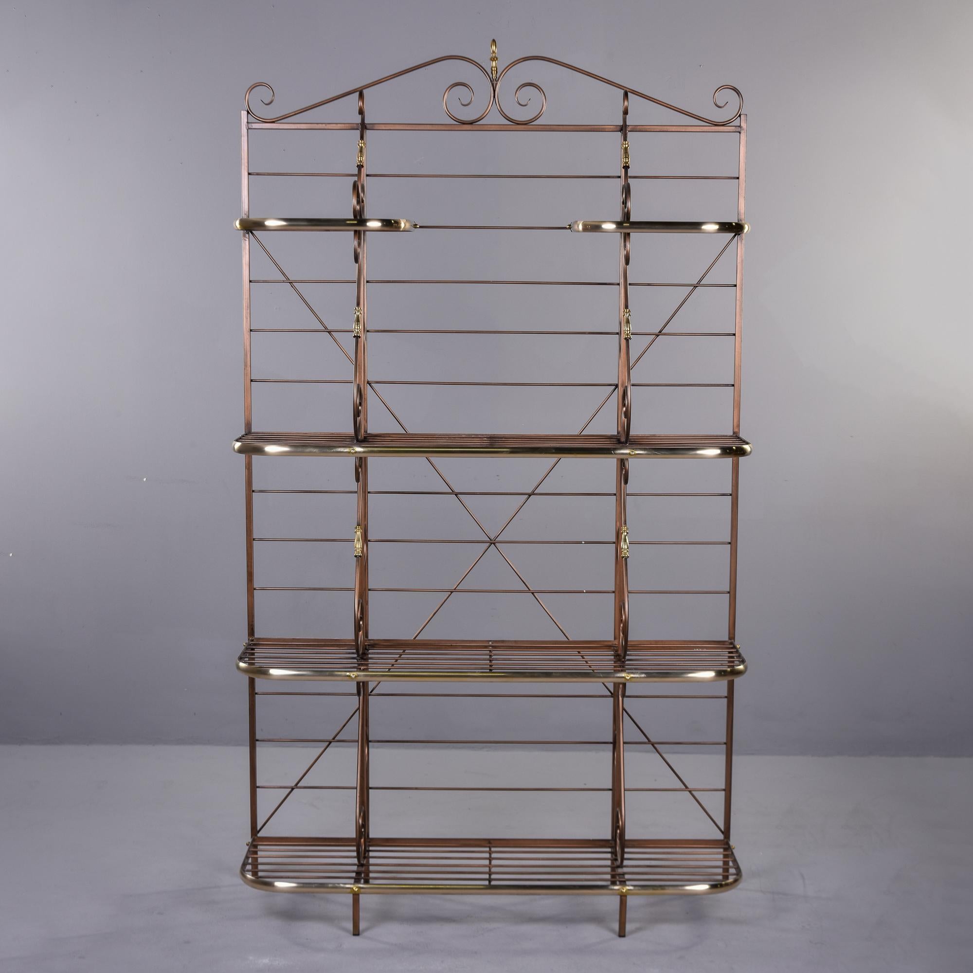 Circa 1980s baker’s rack found in the US has a bronze-colored metal frame with brass bumpers and accents. Five shelves in total: two small ones at the top with three full-width shelves below. Top has a scrolled metal crest. Unknown maker. Very good