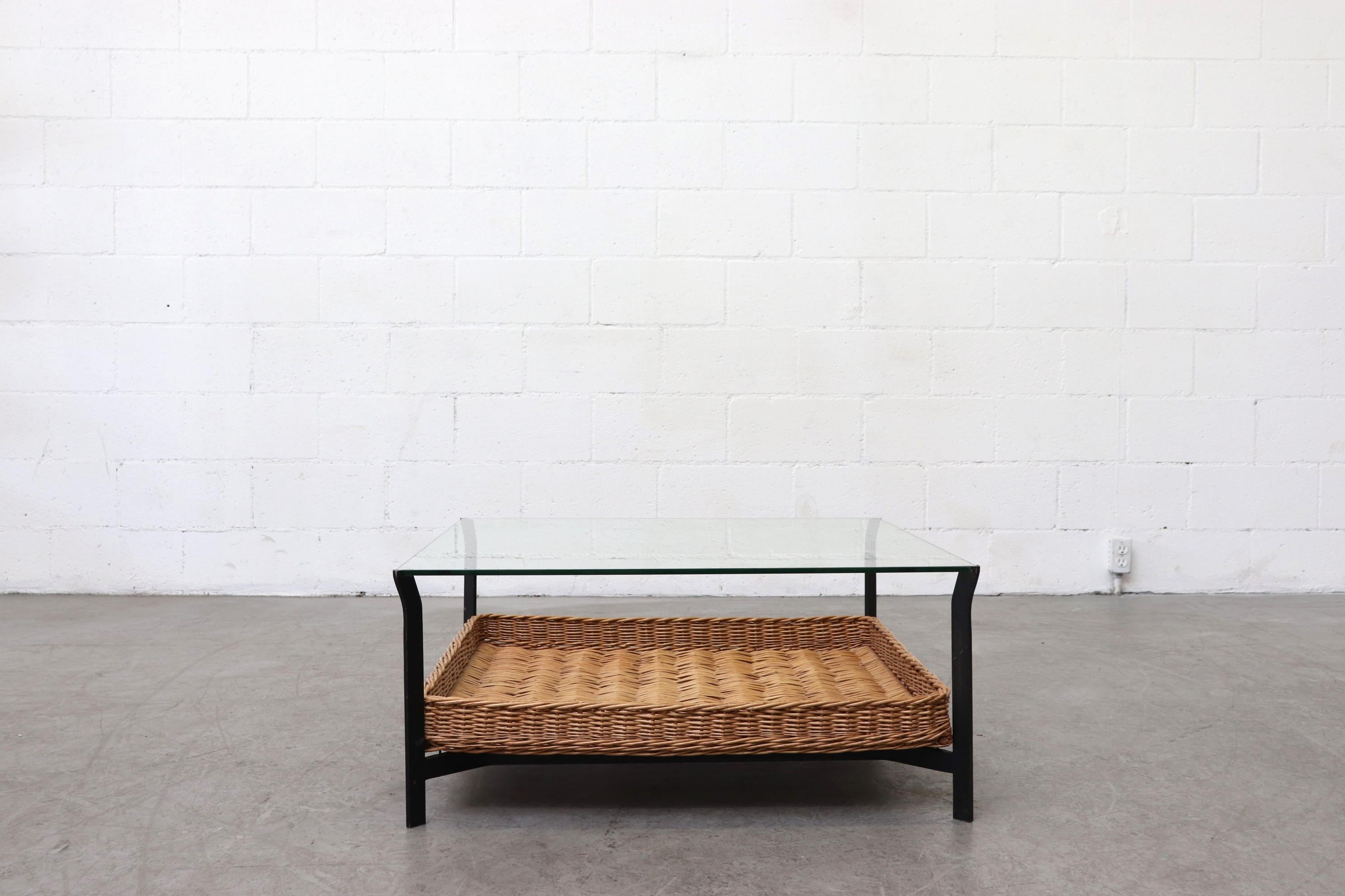 Square two tiered mid-century black enameled metal and glass coffee table with rattan lower basket shelf. In original condition with visible wear to the glass and frame, including some chipping of glass to the corners and minimal wear to the rattan