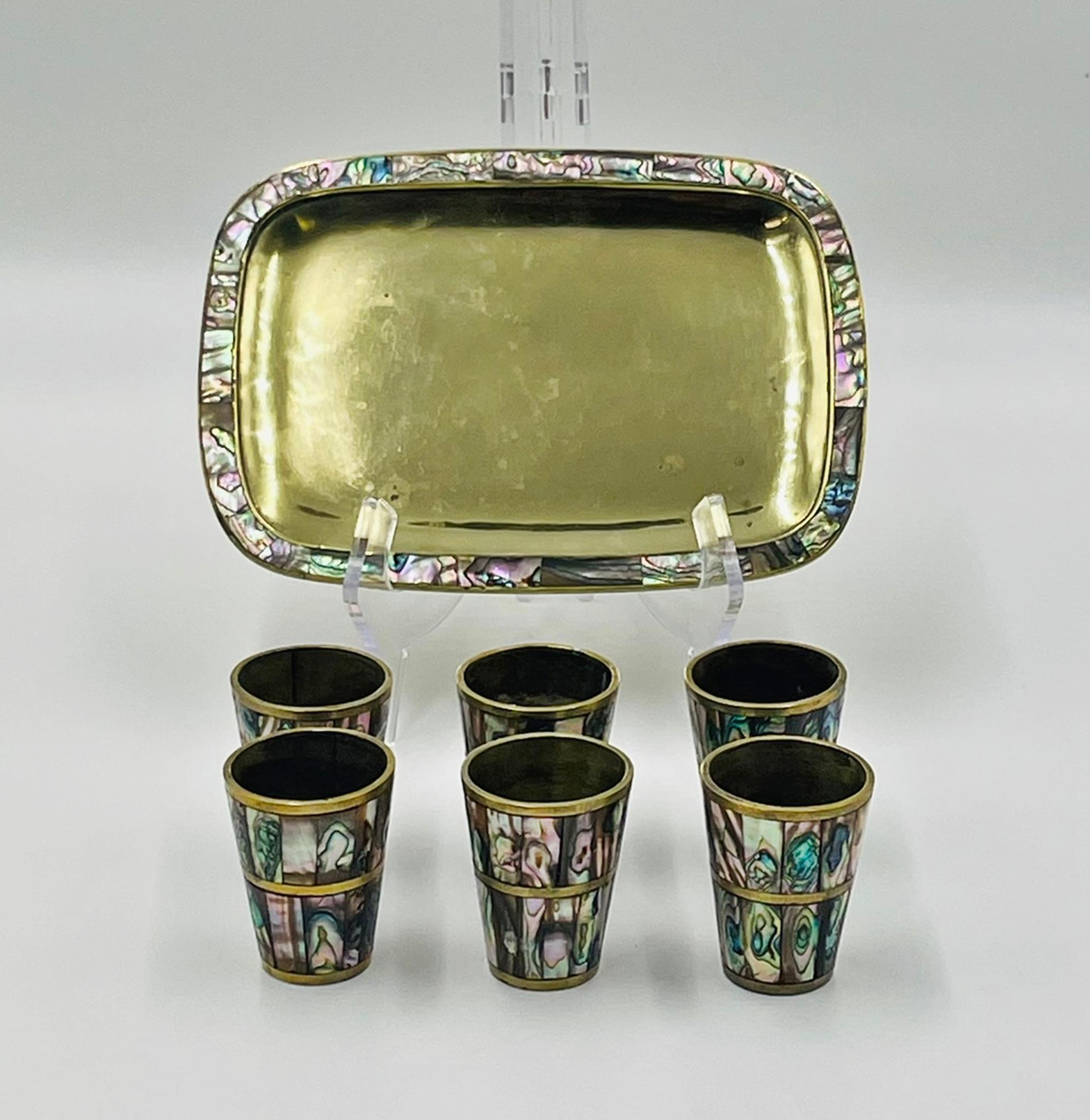 Elegante juego de bar mexicano de mediados de siglo compuesto por seis vasos de chupito de latón y una bandeja rectangular a juego, cada uno adornado con concha de abulón meticulosamente incrustada a mano. Las baldosas de nácar iridiscente brillan