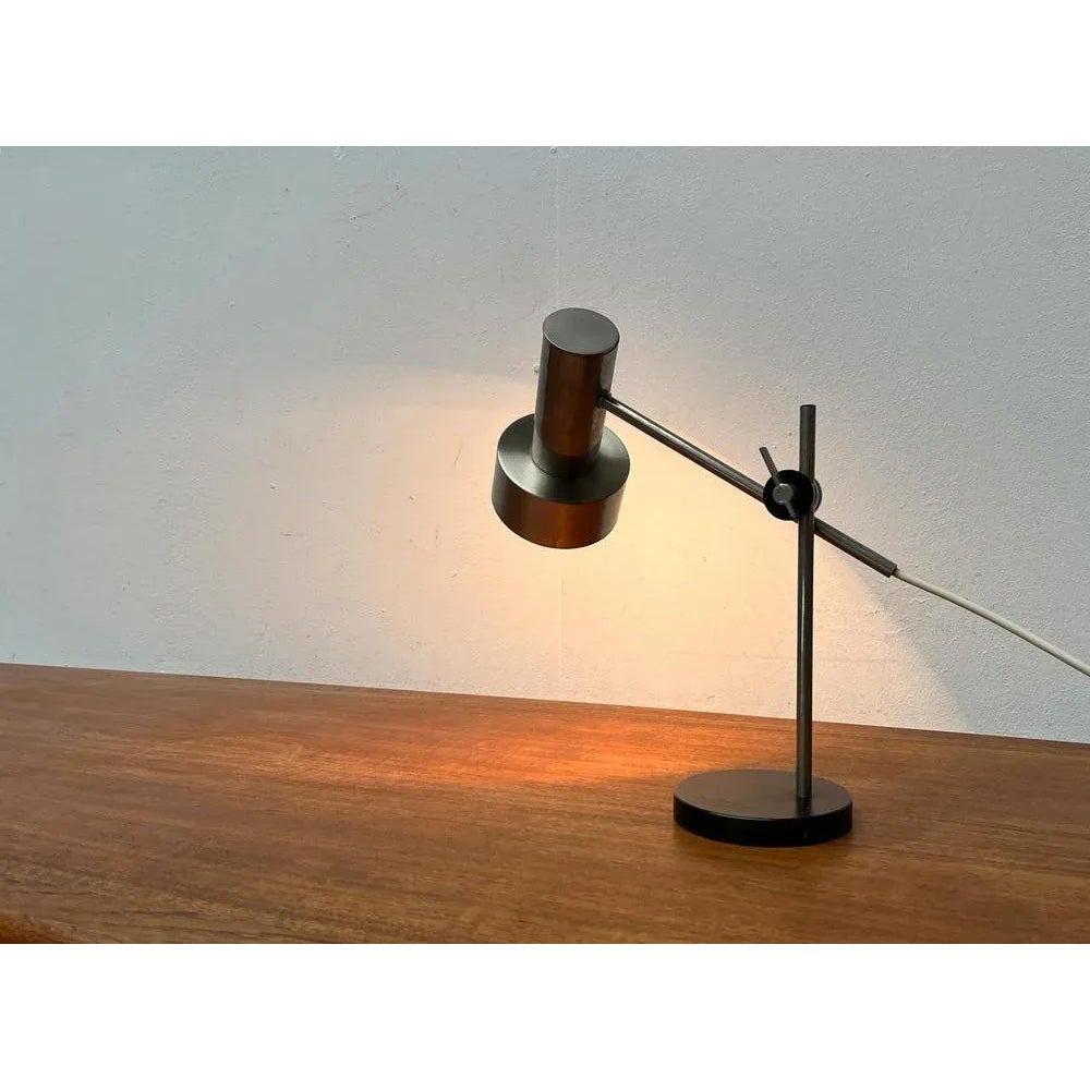 Pequeña lámpara de sobremesa de mediados de siglo.

Llama la atención la forma de radiador minimalista del cabezal de la luz. Dos cilindros simples de distinto tamaño. El cabezal de la luminaria se asienta en el extremo de un brazo que está unido al