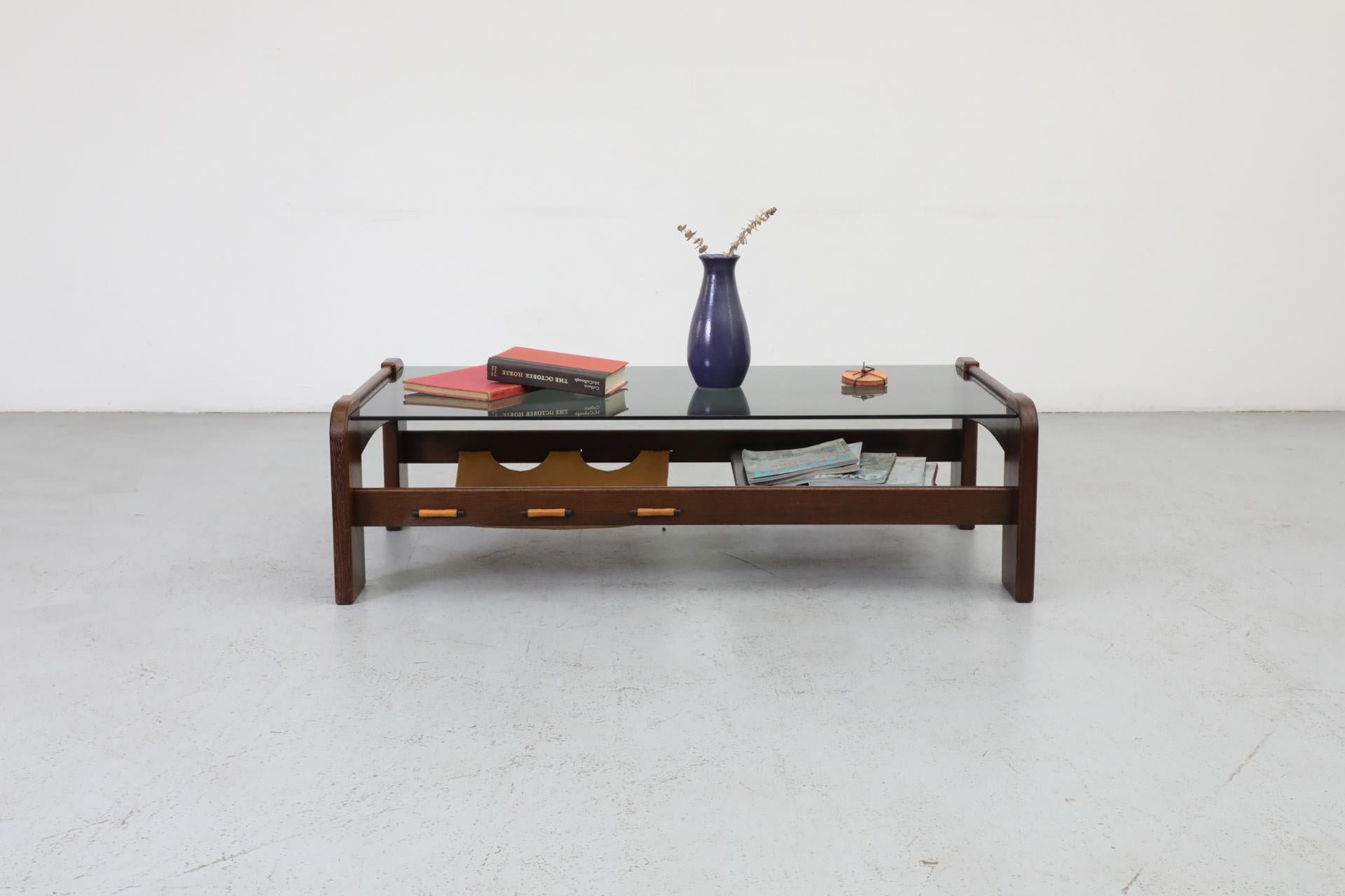 Charmante table basse hollandaise du milieu du siècle dernier des années 1970, avec un cadre en bois wengé, un plateau en verre fumé, une étagère inférieure en lattes et un porte-revues en cuir fauve. En état original avec une usure visible