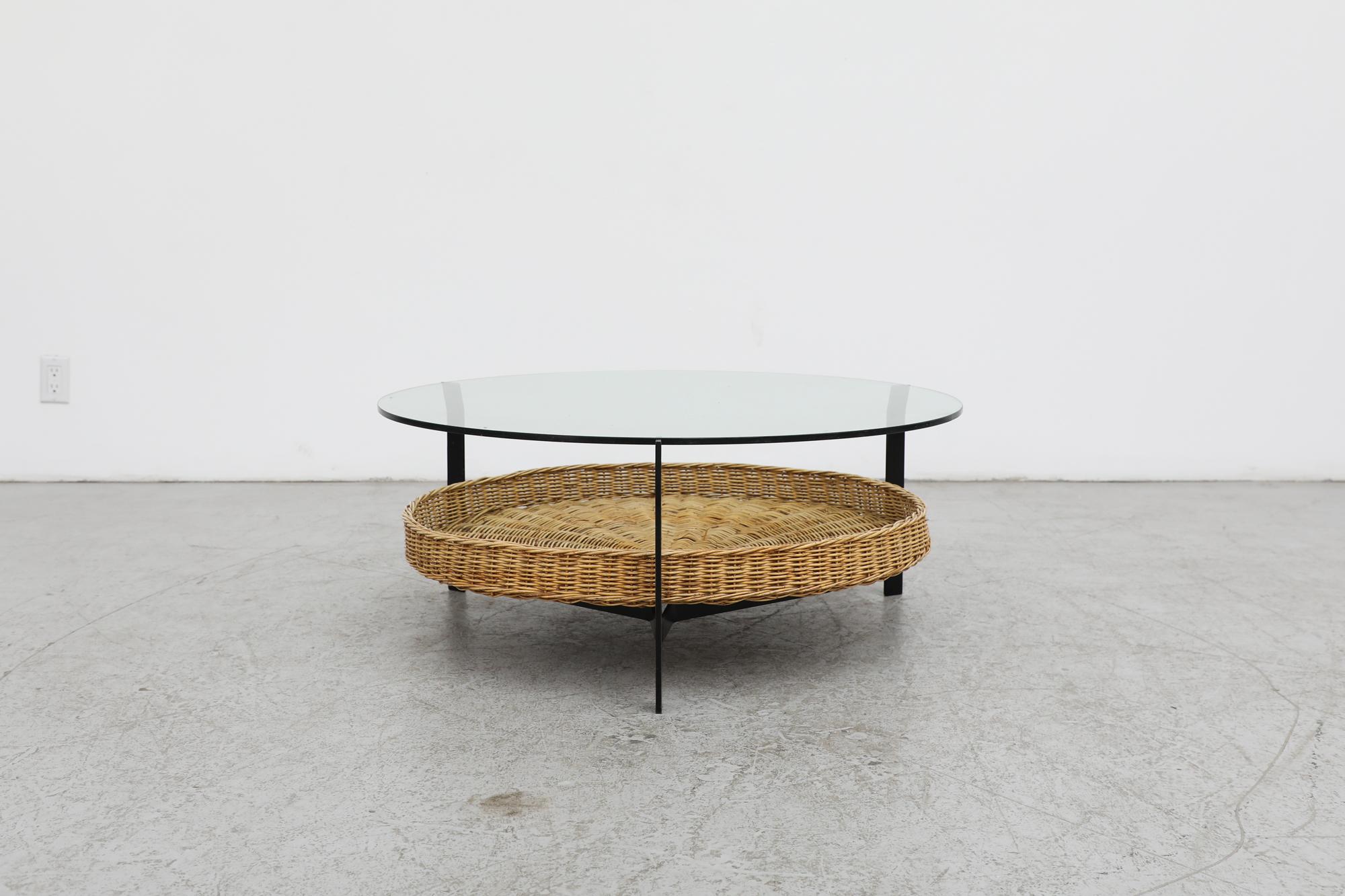 Mid-Century, Rudolf Wolf designed round glass coffee table with woven wicker basket and black enameled metal frame. The flat black steel frame curves slightly outwards to hold the glass. The glass shows visible wear and scratching. In original