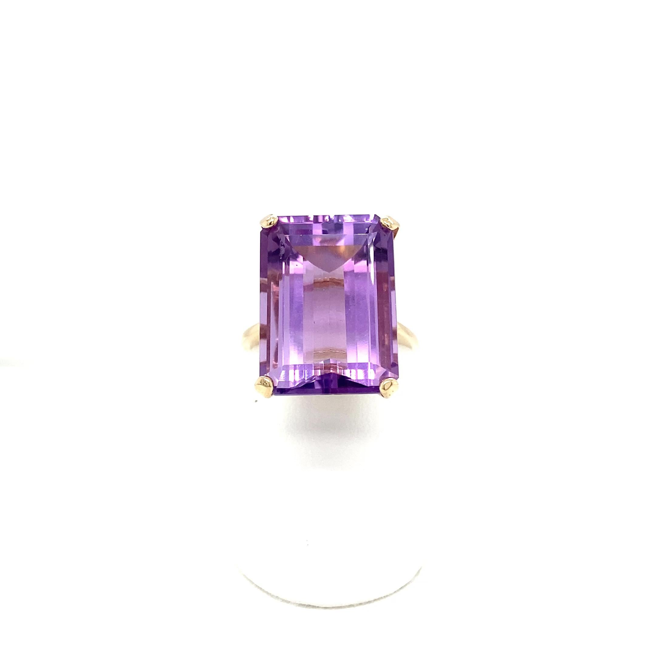 Questo anello da cocktail della metà del secolo scorso presenta un'ametista solitaria dal taglio smeraldo incastonata in una classica montatura a quattro punte in oro giallo 14 carati lucidato a specchio.  La gemma centrale di colore viola reale