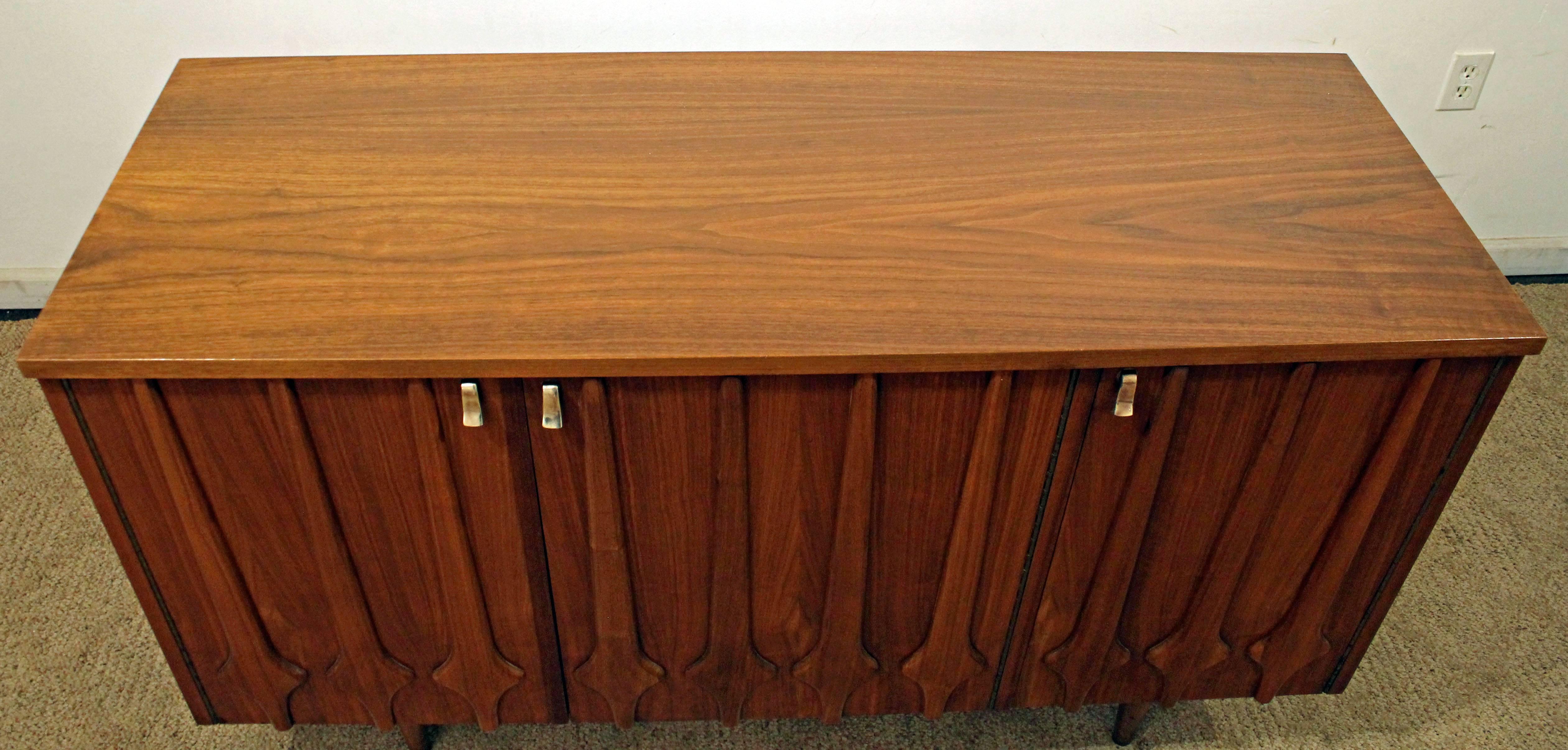 Offered is a walnut credenza featuring 3 doors, shelving, and brass pulls. 