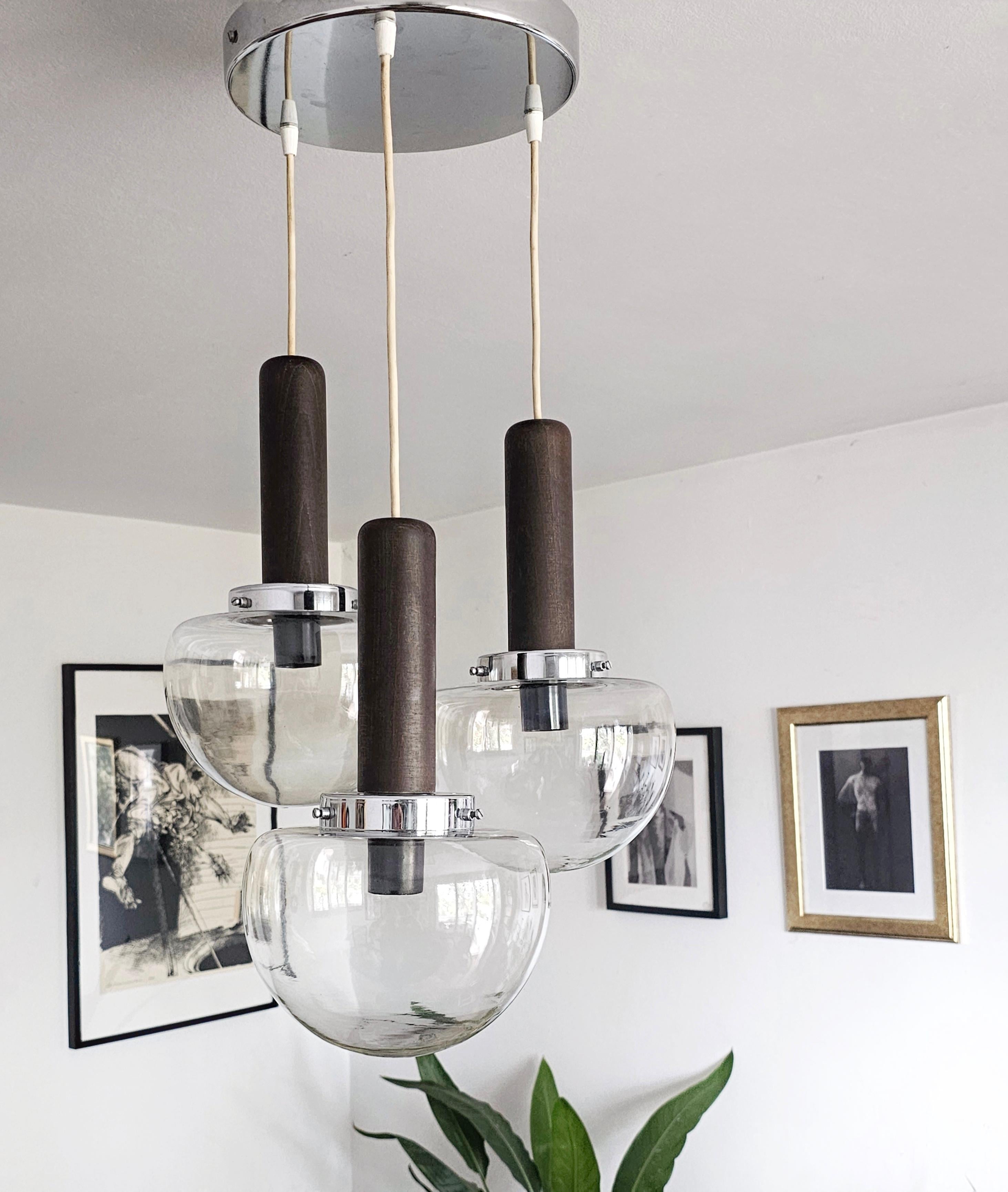 In this listing you sill find a gorgeous minimalist cascade chandelier manufactured by Sijaj Hrastnik. It features three levels, consisting of wooden cylinders and lightly smoked glass shades shaped as mushroom hats, with the circular chrome