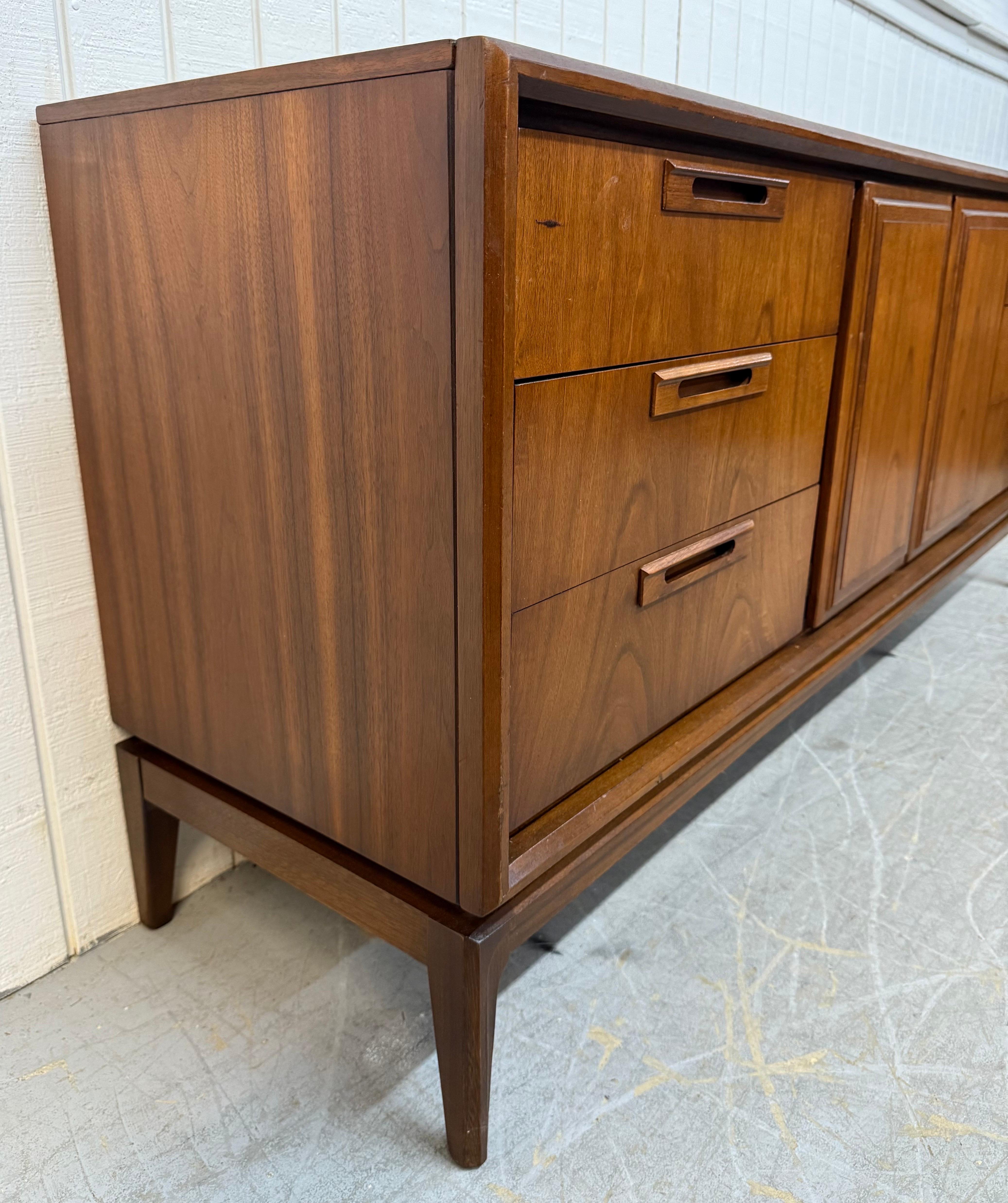 Cette annonce concerne une commode en noyer à 9 tiroirs de style moderne du milieu du siècle. Elle se caractérise par un design rectiligne, trois tiroirs à chaque extrémité avec des poignées en bois encastrées, des portes centrales qui s'ouvrent sur