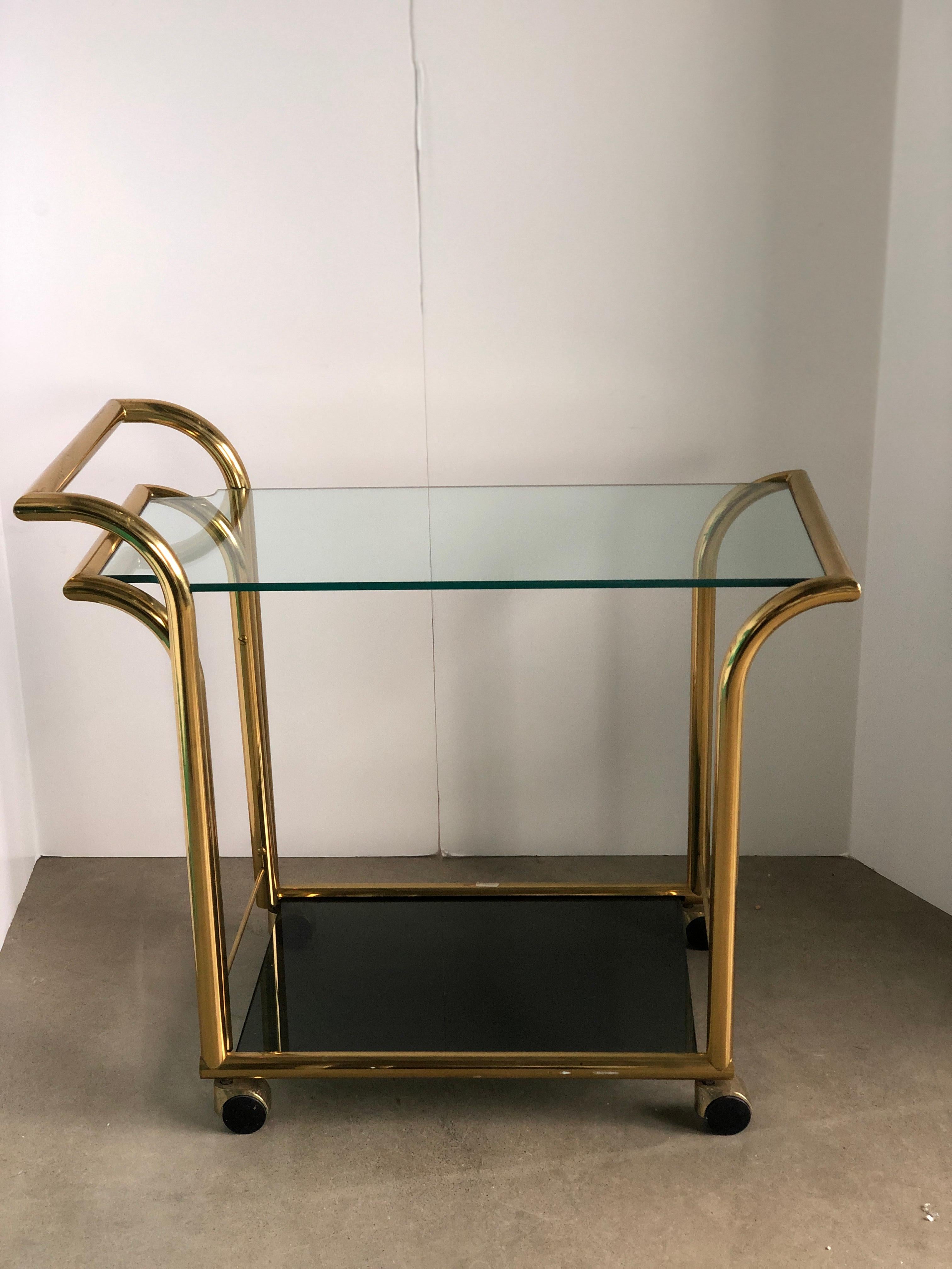 Offered is a Mid-Century Modern Art Deco style, two-tier clear glass on top and smoked glass on bottom with a brass frame and wheel canopy bar cart or service trolley signed by Design Institute America Italy. Both glass shelves sit secured in the