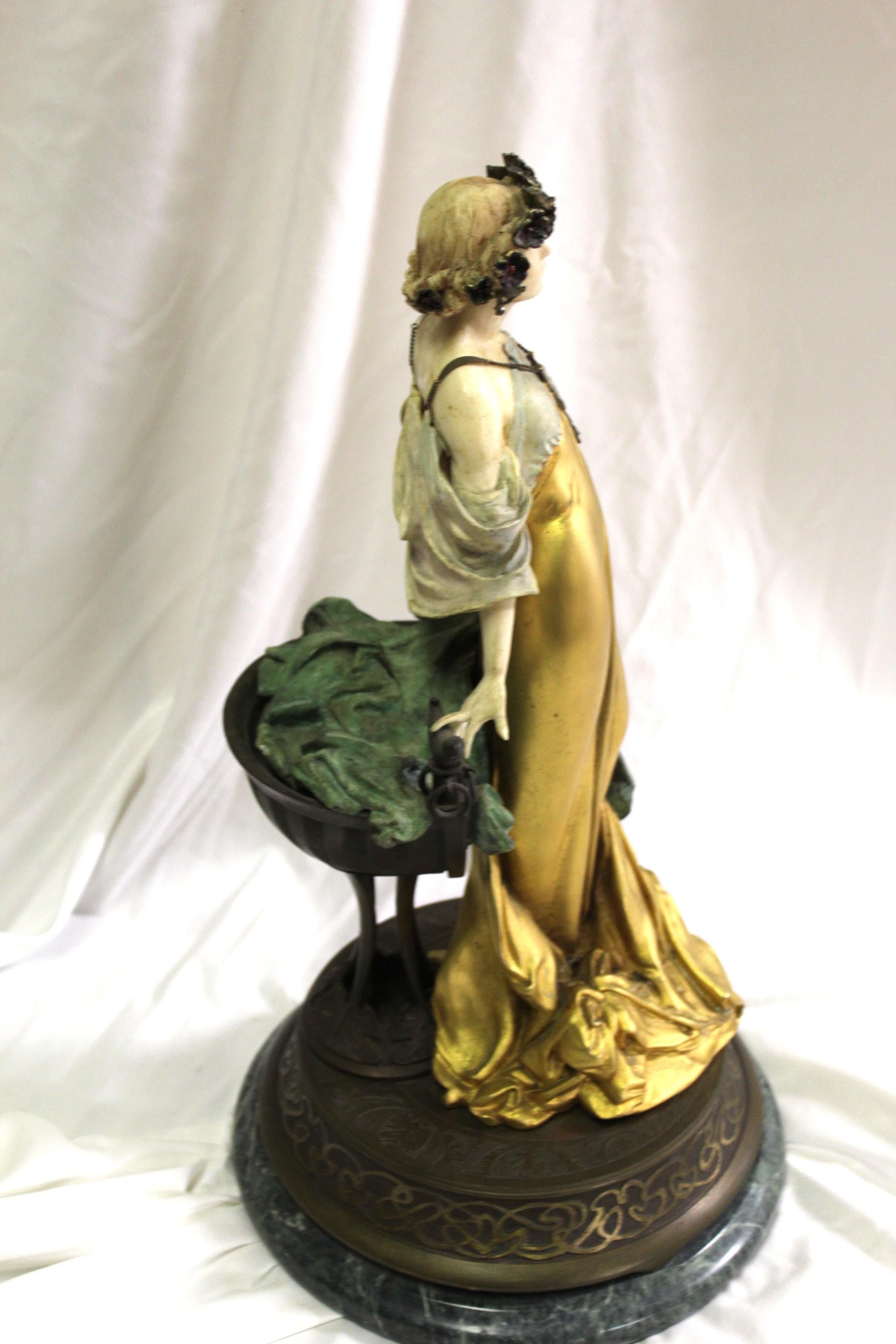 Questa raffinata scultura in bronzo ritrae una graziosa figura femminile in stile Art Nouveau, che si erge sicura con un'espressione serena. Indossa un fluente abito dorato che aderisce elegantemente alle sue forme, accentuando la bellezza