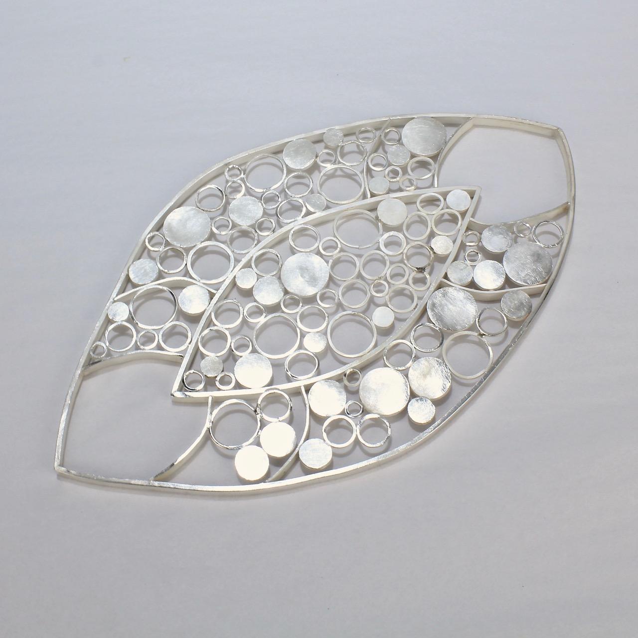 A whimsical, modernist sterling silver trivet. 

With a hand soldered ring and dot pattern in an asymmetrical oval form and with a brushed finish.

Perfect for a wine bottle or serving bowl!

The reverse is stamped Sterling and with the artist's
