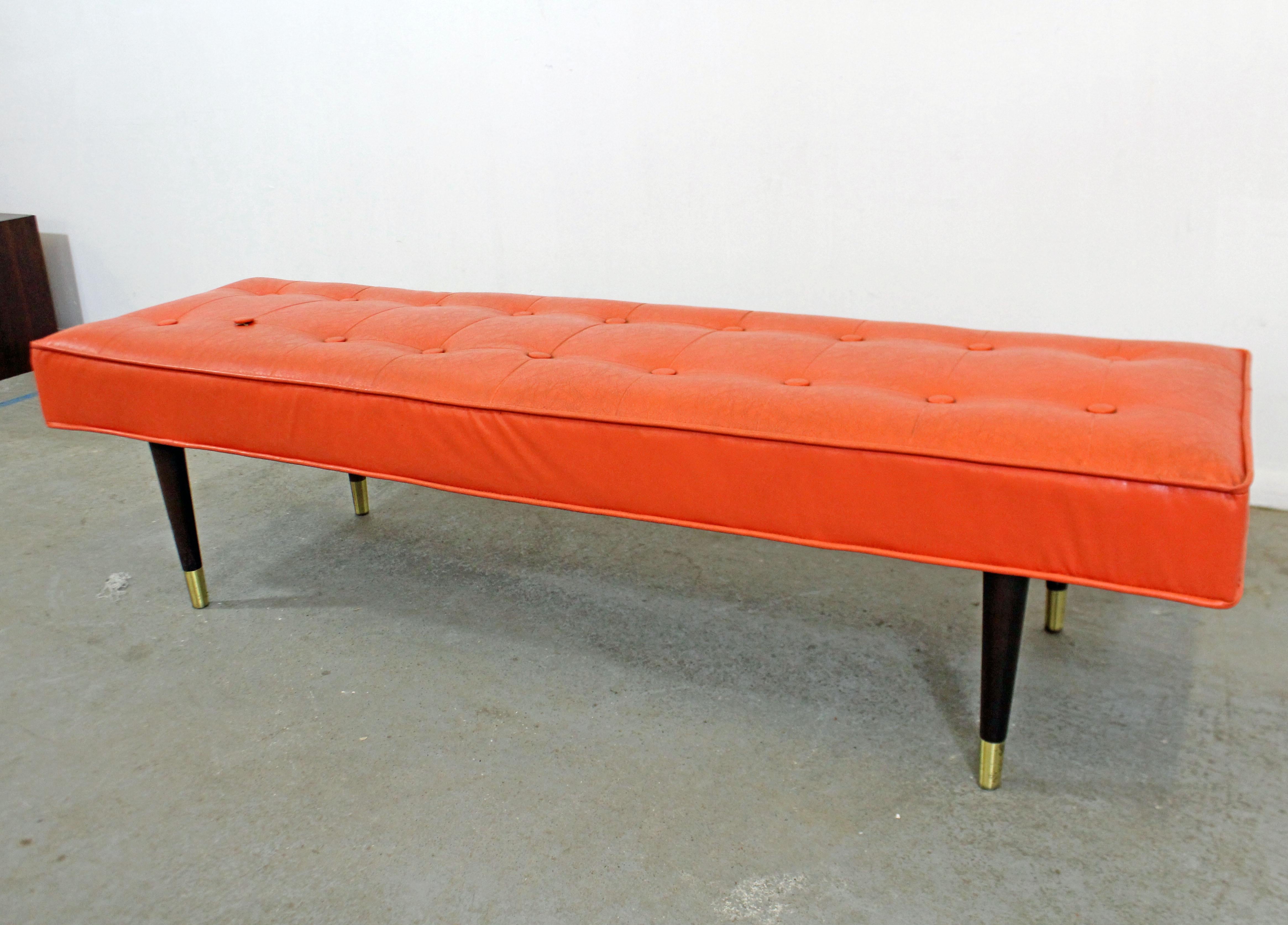 Offered is a mid-century modern bench featuring atomic orange vinyl upholstery and walnut pencil legs with brass capped feet. It is in decent, structurally sound condition with normal age wear. Needs some restoration (see condition and photos). It