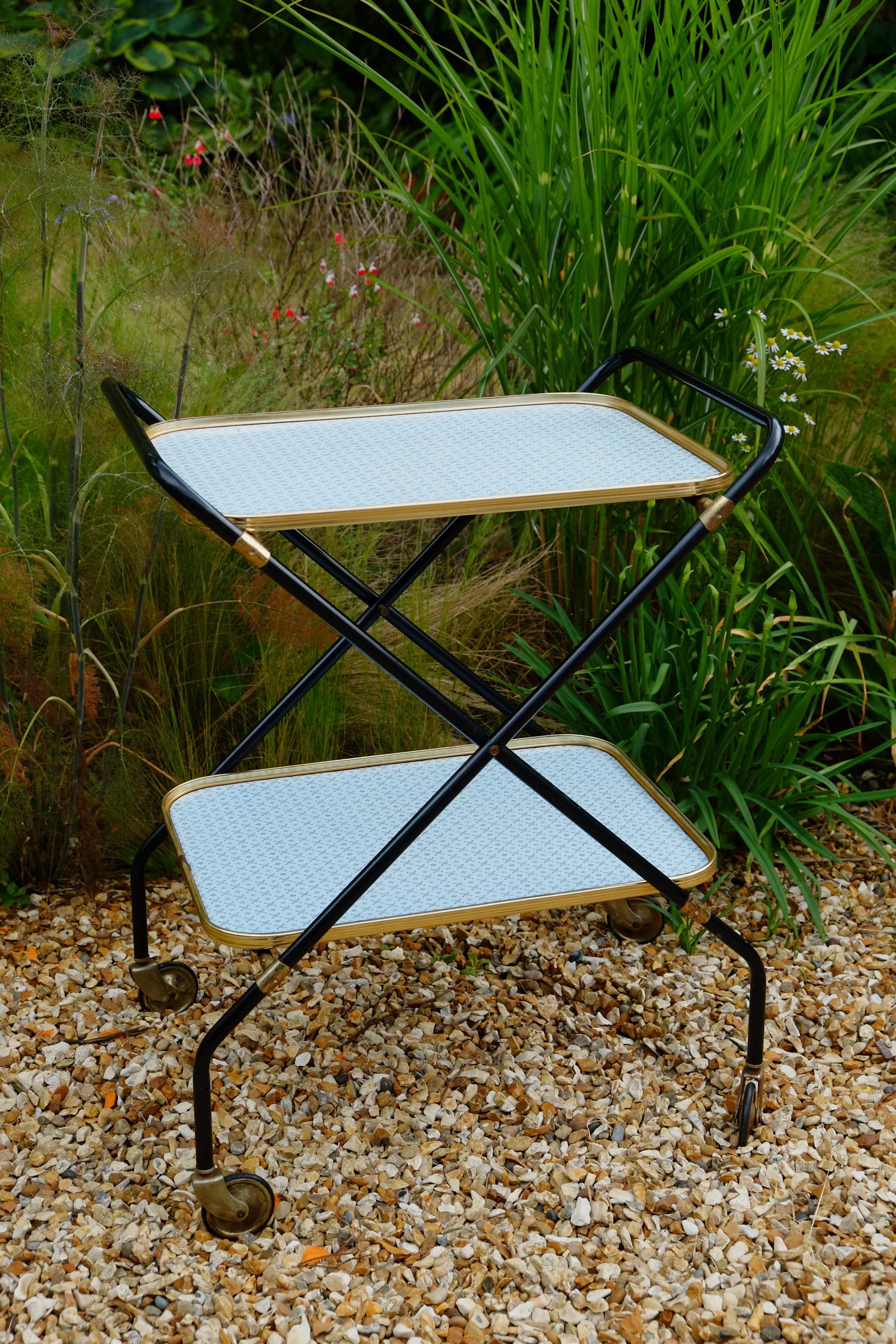 A rare Mid-Century Modern folding trolley bar cart from the 1950s or 1960s. The trolley has a gorgeous angular X shaped leg structure with large brass coloured wheels. Two beautiful gold lipped blue laminate trays with an elegant pattern across the
