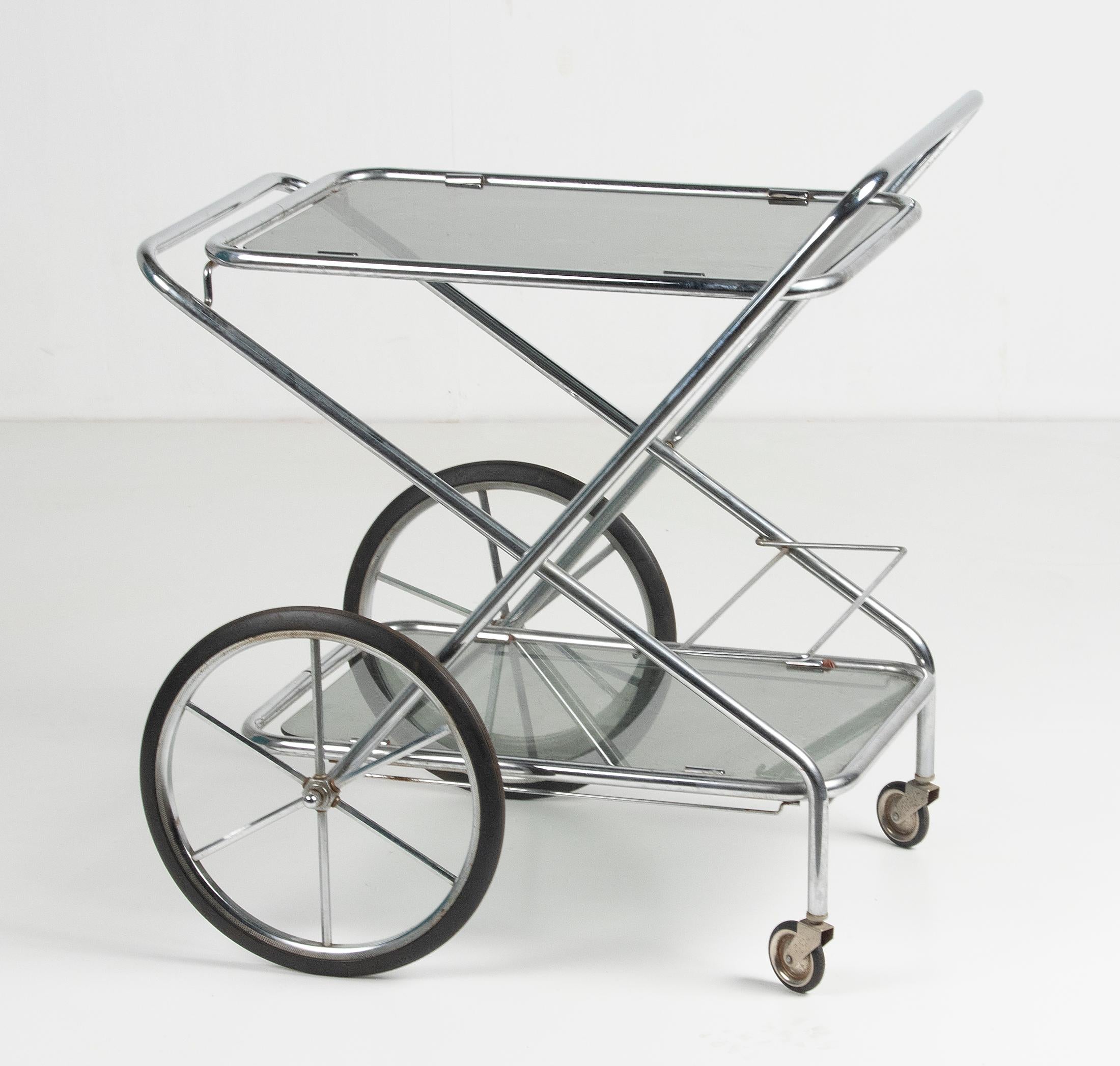 A mid century folding bar cart made of polished chrome-plated iron. It has two smoked glass tops, swivel wheels and a rack for bottle. It's made in Belgium, circa 1960-1970. The cart is foldable. Rust stains on the small swivel wheels.
 