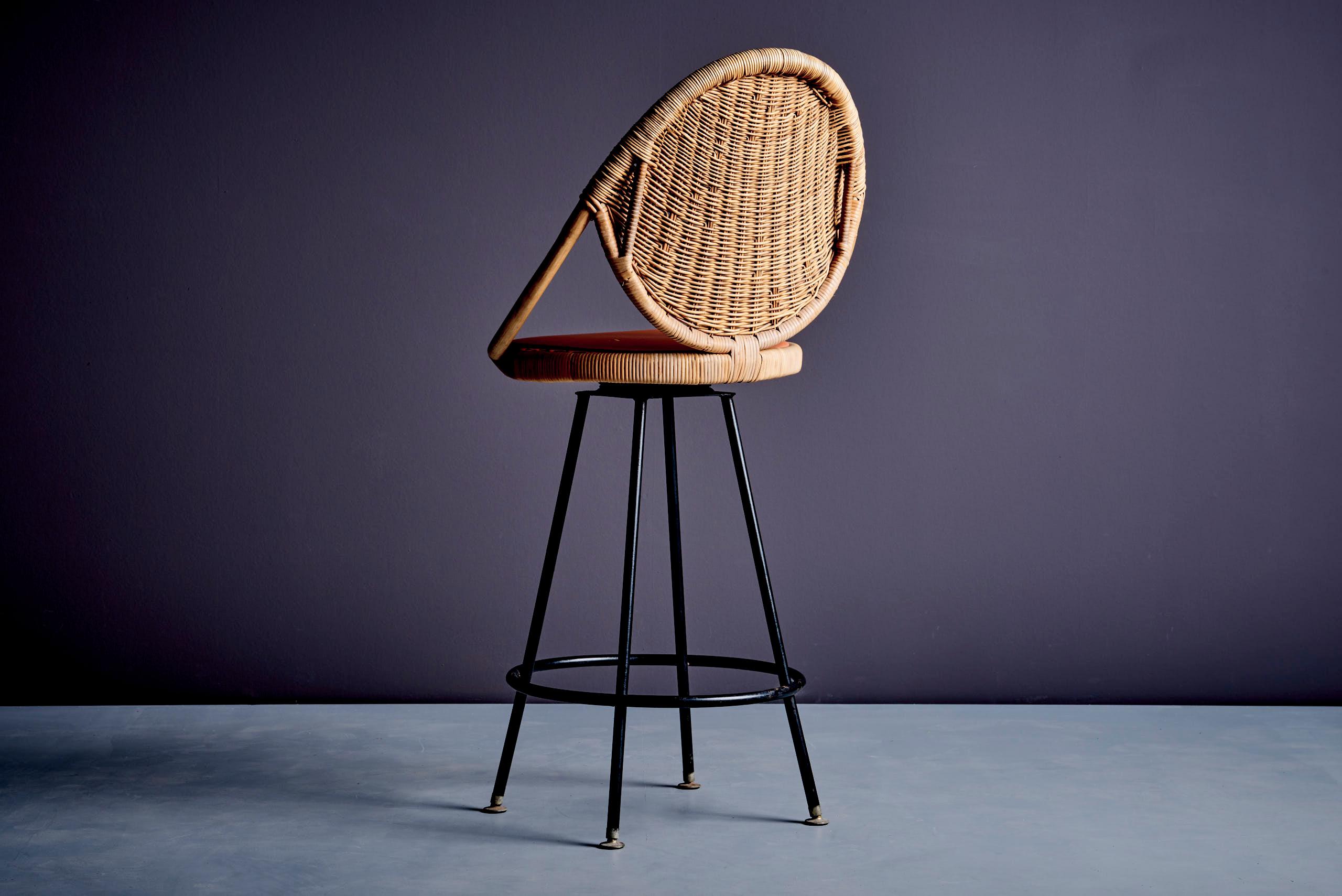 This elegant bar stool designed by Danny Ho Fong, featuring a round rattan-wrapped backrest and seat on a sleek black wrought iron base. The stool has a sculptural form, with curved arms integrated into the wicker construction. The frame includes a