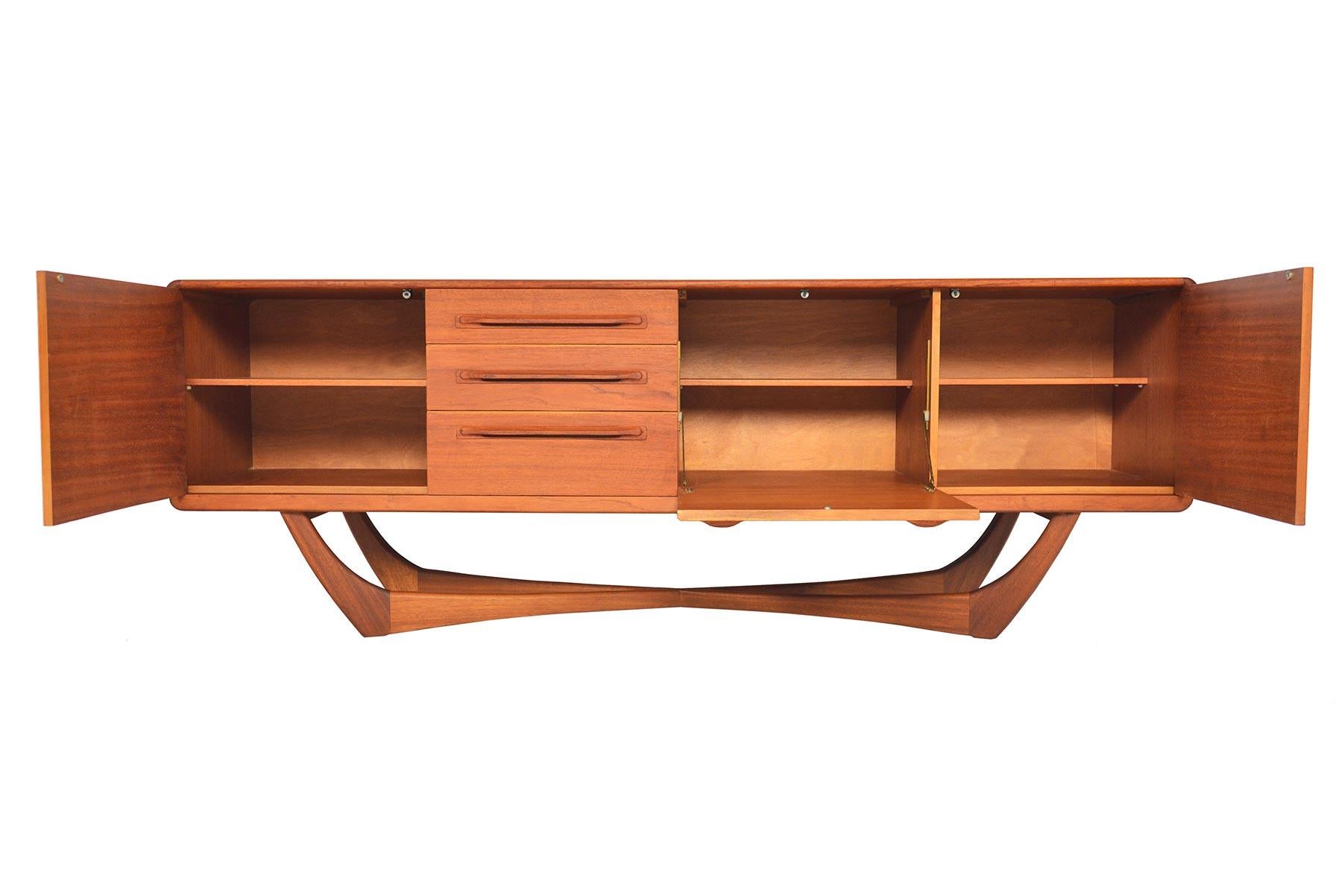 This exceptional Scottish modern teak credenza was designed by Val Rossi for Beithcraft. The case sits on a dramatic parabolic x- form base. The left and right bays open to reveal a removable shelf. The center bar drops down to provide a serving