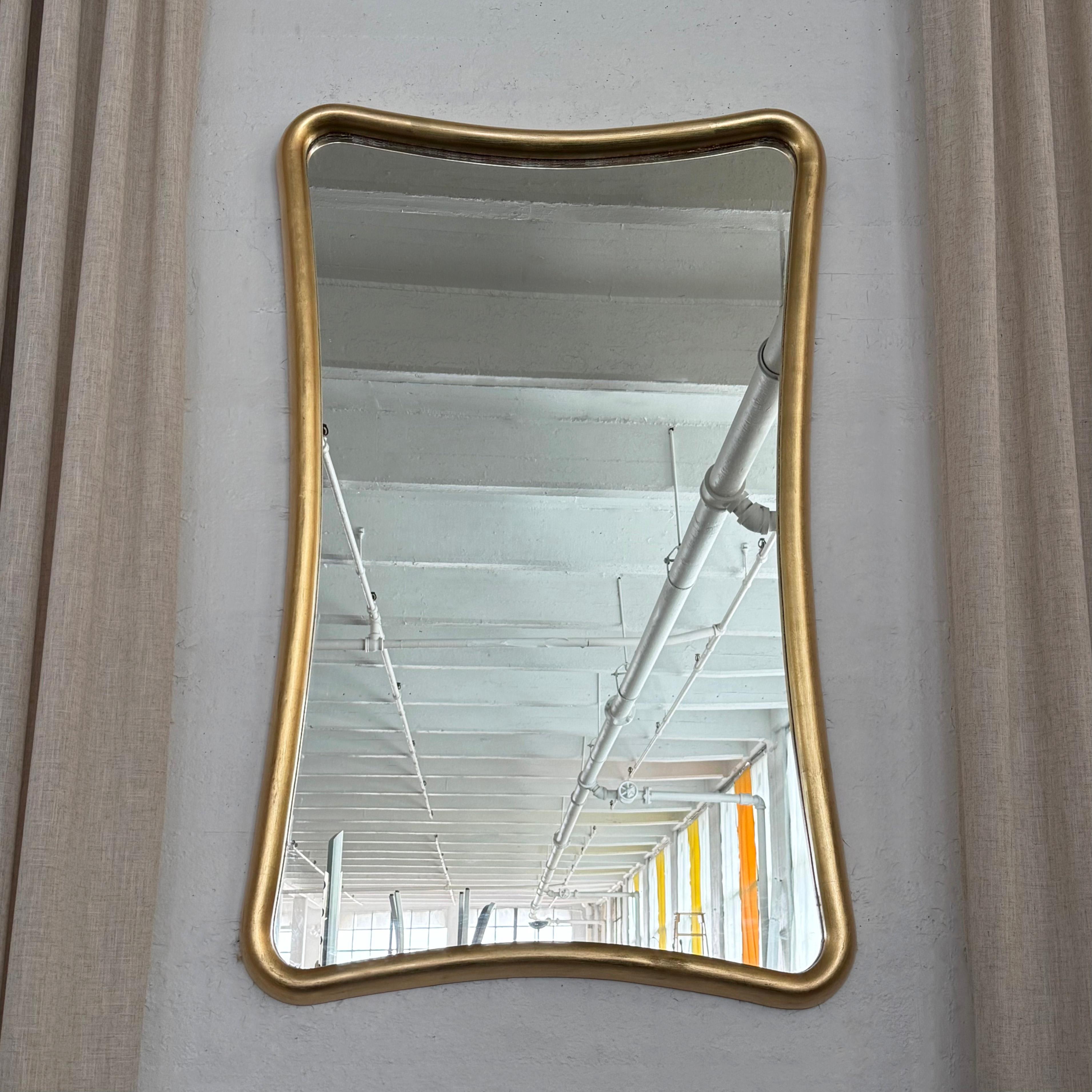 Ce miroir mural doré de style régence hollywoodienne et de facture soignée présente un cadre en bois épais de forme organique, aux lignes élégantes, aux angles arrondis et au dos en bois massif. La finition dorée brille tout en présentant une belle