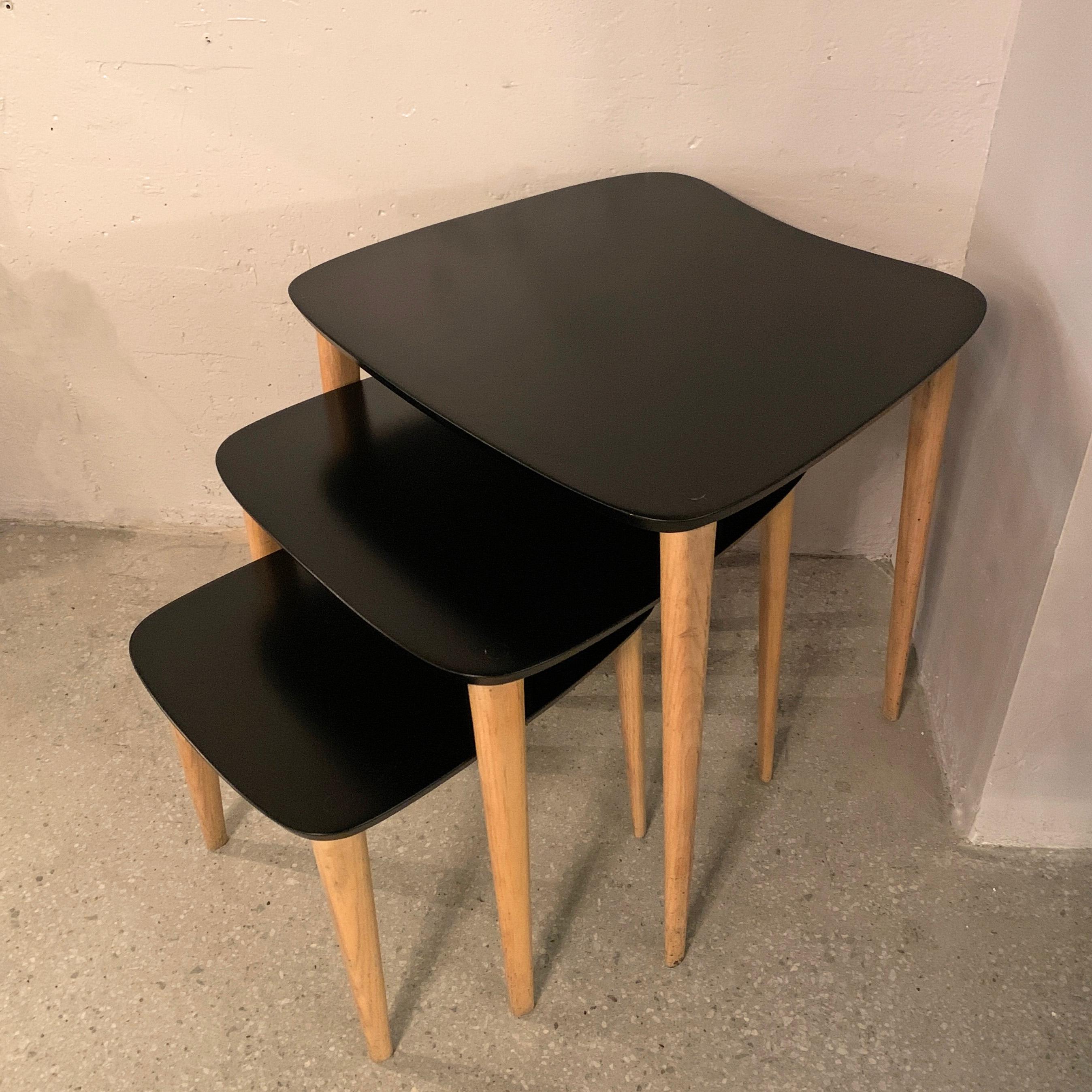 Trio of Mid-Century Modern, nesting tables feature biomorphic, black lacquered tops with contrasting, tapered natural legs. The middle table measures: 19 inches width x 19.5 inches depth x 19.25 inches height and the bottom table measures: 14.5