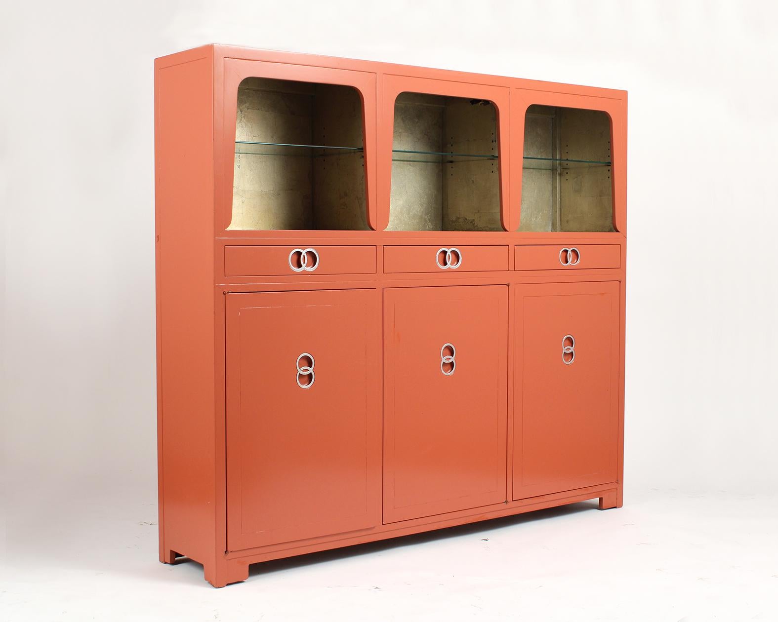 This Mid-Century Modern bookcase by Baker is in good condition and has its original orange painted finish. The top section has three spaces each with an adjustable glass shelf, and original gold painted interior. Below that there are three pull out
