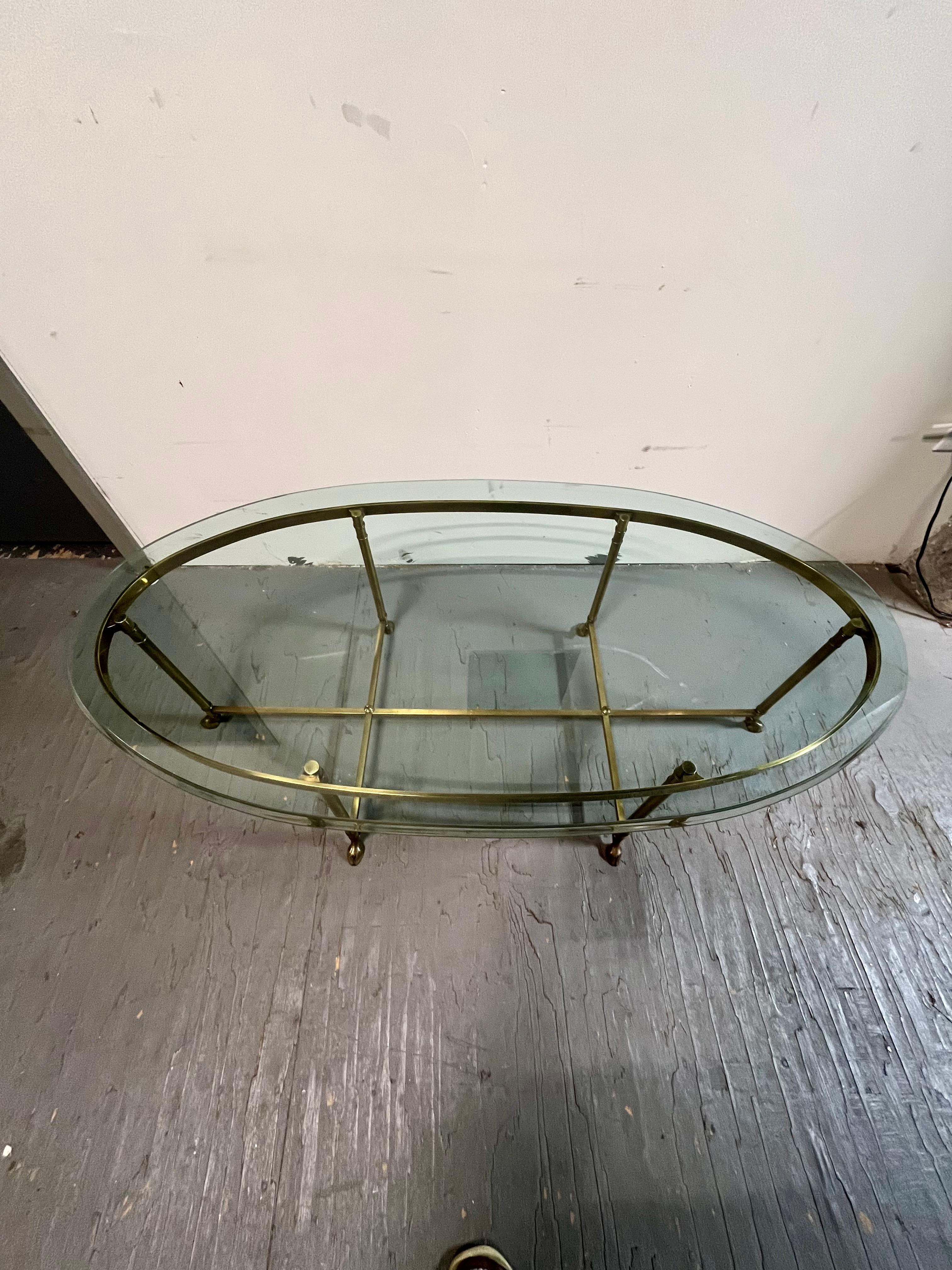 Cette table à cocktail ovale de style moderne du milieu du siècle a été créée à la manière de Jansen, vers 1960. La table a un plateau en verre biseauté de forme ovale, soutenu par six pieds en laiton élancés. Chaque pied repose sur un sabot stylisé