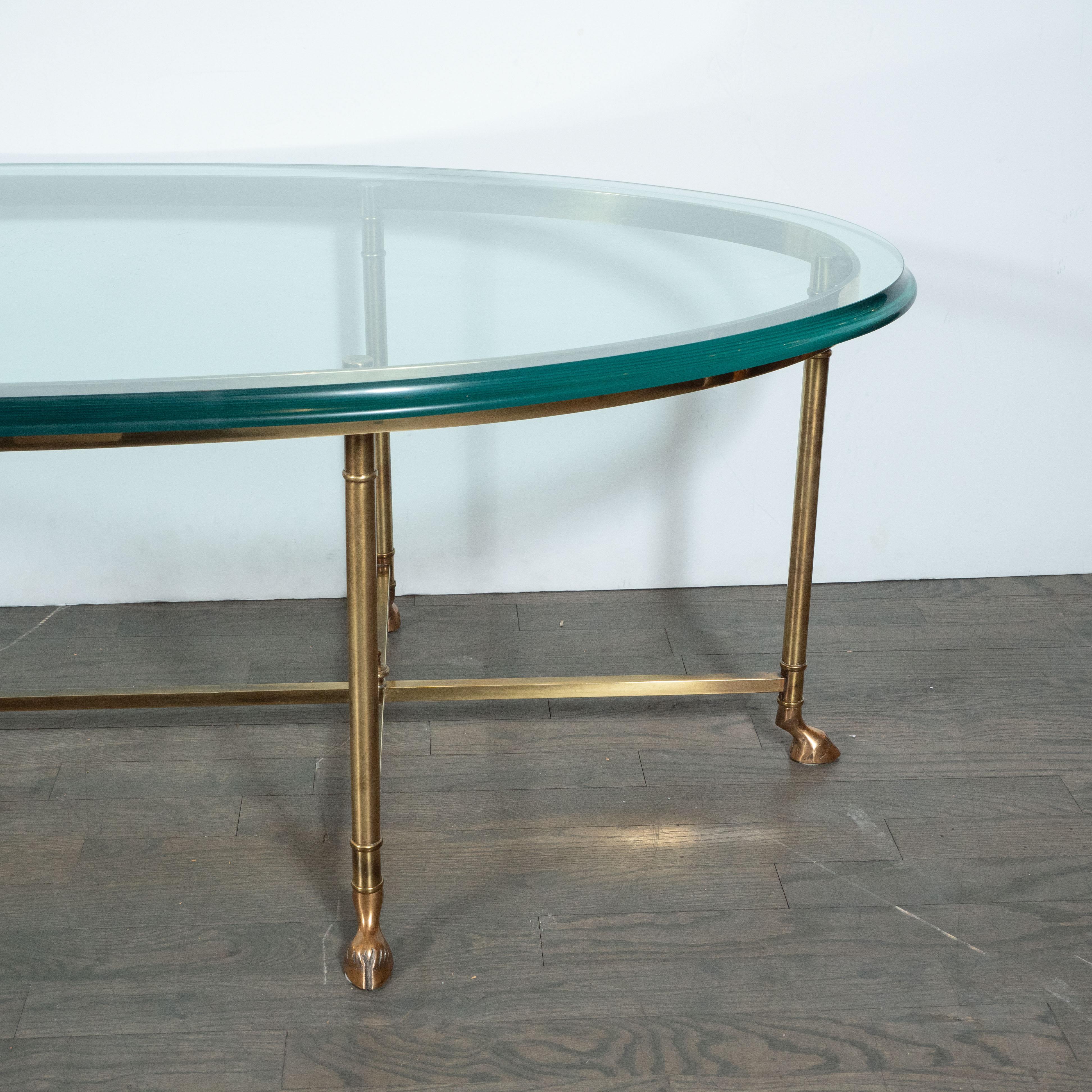 This Mid-Century Modern oval cocktail table was created in the manner of Jansen, circa 1960. The table has an oval-shaped bevelled glass top which is supported by six slender brass legs. Each leg sits on a stylized brass hoof, a nod to classical
