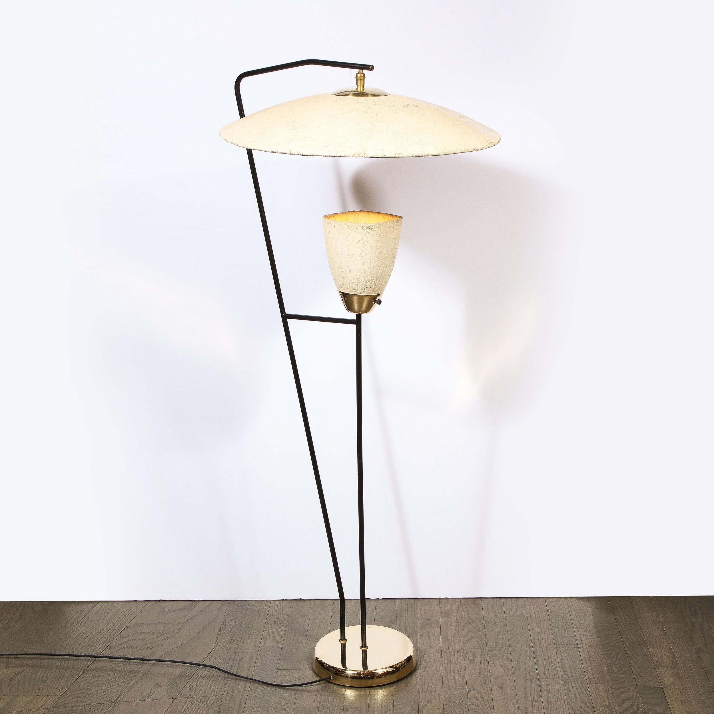 This sophisticated Mid-Century Modern floor lamp was realized in the United States circa 1950. It features a circular button-like base in polished brass from which two cylindrical supports in black enamel ascend. One of the supports rises from the