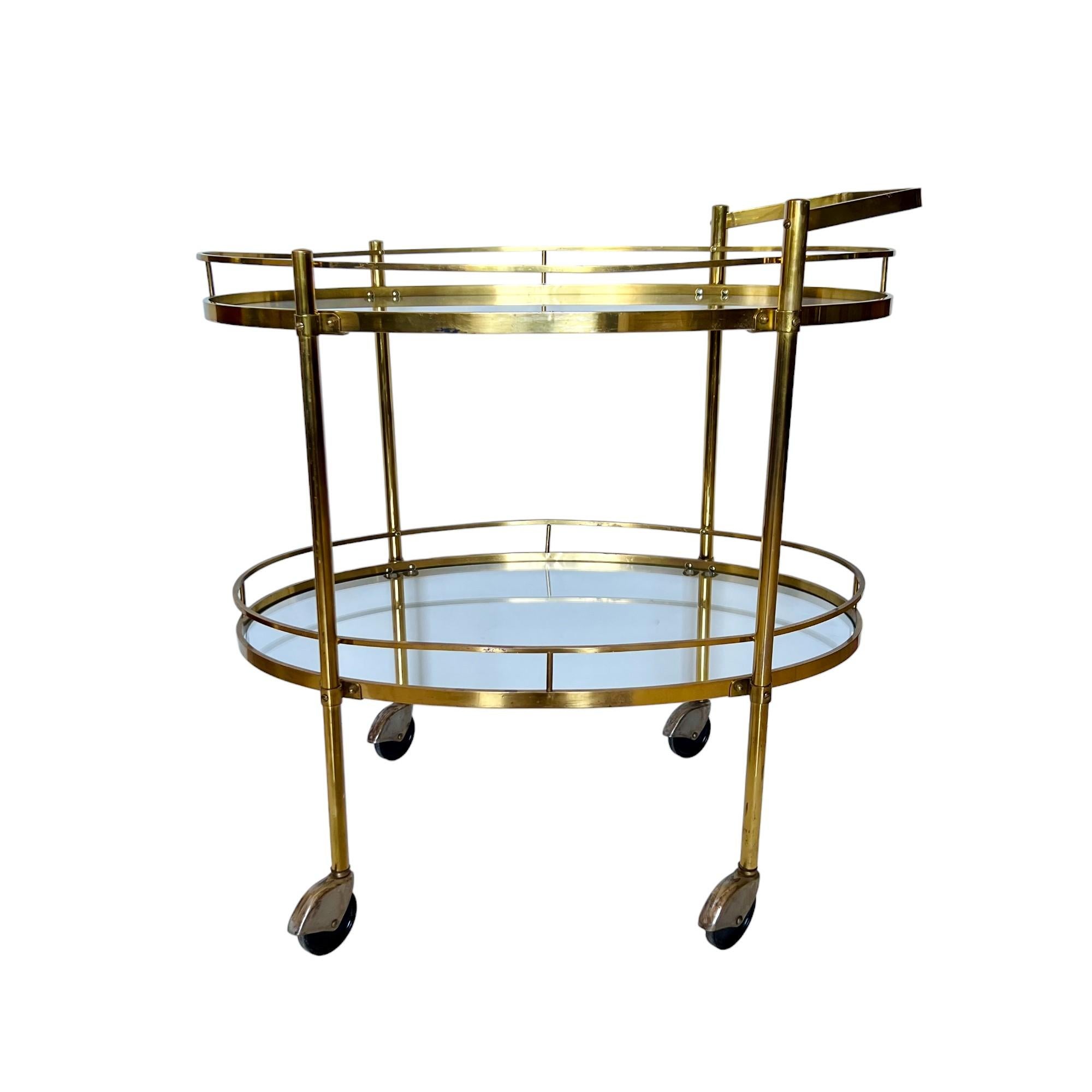A vintage 1960's Hollywood Regency style two tier brass and glass oval bar or tea cart. It features a solid brass frame with an angular handle, floating form transparent glass shelves and black rolling casters with steel wheel covers. 

Dimensions: