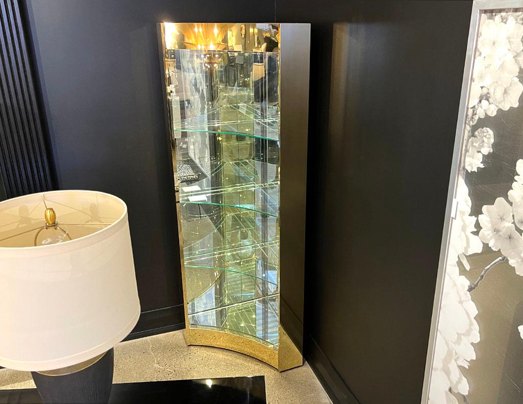 Mid-Century Modern brass and glass corner curio display cabinet. All original from USA, 1970’s. Featuring unique curved brass frame with mirror back and glass shelves. Cabinet features interior top light. Price includes complimentary curb side