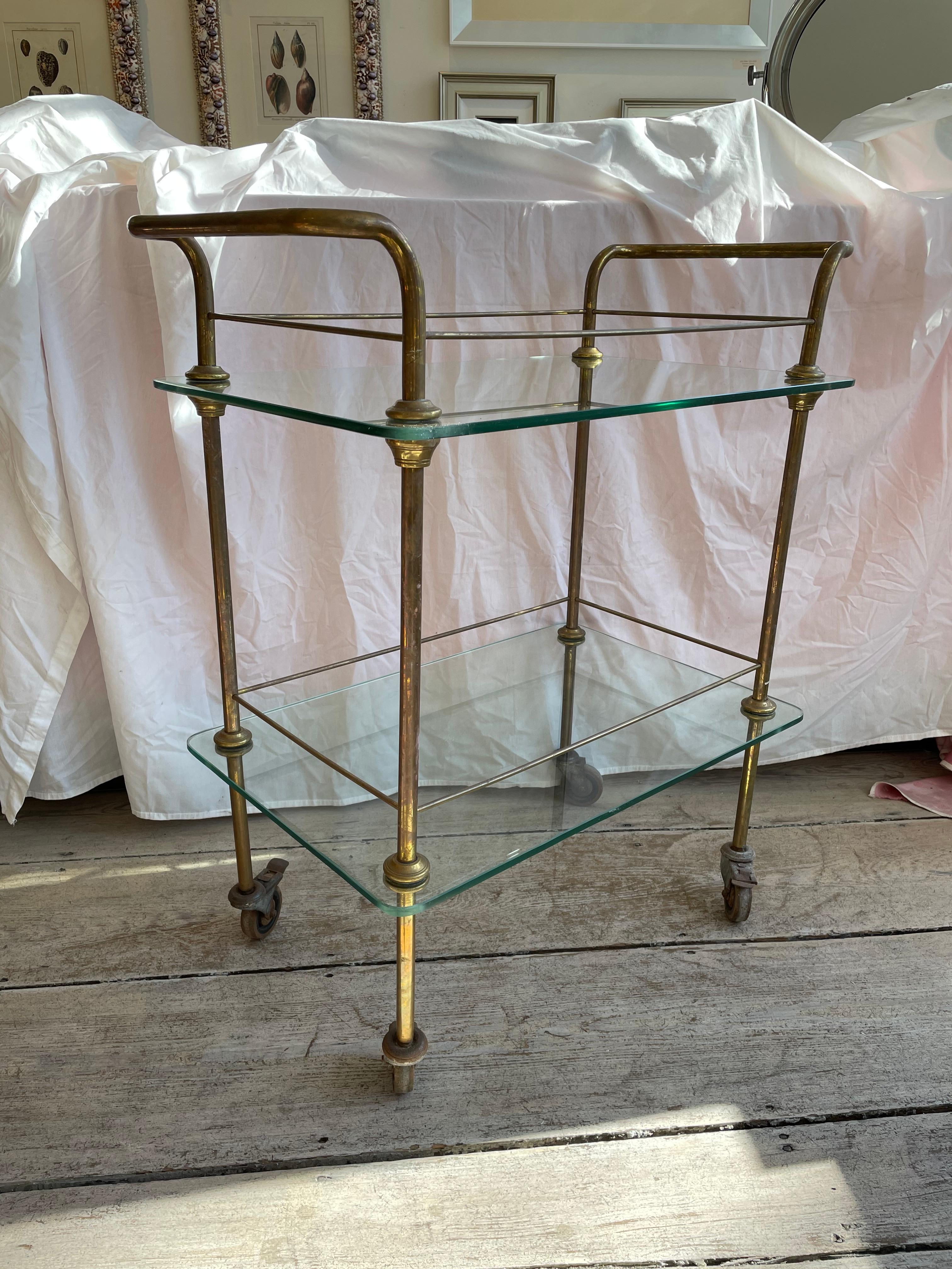 A Mid-Century Modern brass bar cart. Double sided handles with brass rail around both top and bottom glass shelves. Original wheels complete with brakes. C, 1960's. Glass replaced. Rolls smoothly and easily.