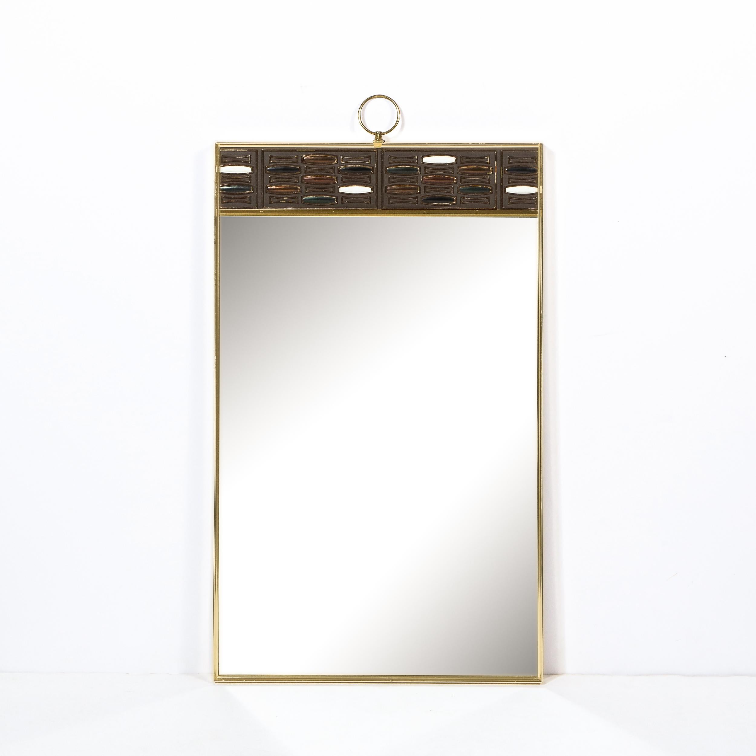 This stunning Mid-Century Modern brass wall mirror was realized in the United States, circa 1960. The mirror offers a rectangular body in lustrous brass with a band of abstract detailing at the top offering stylized ovoid forms in hues of white and