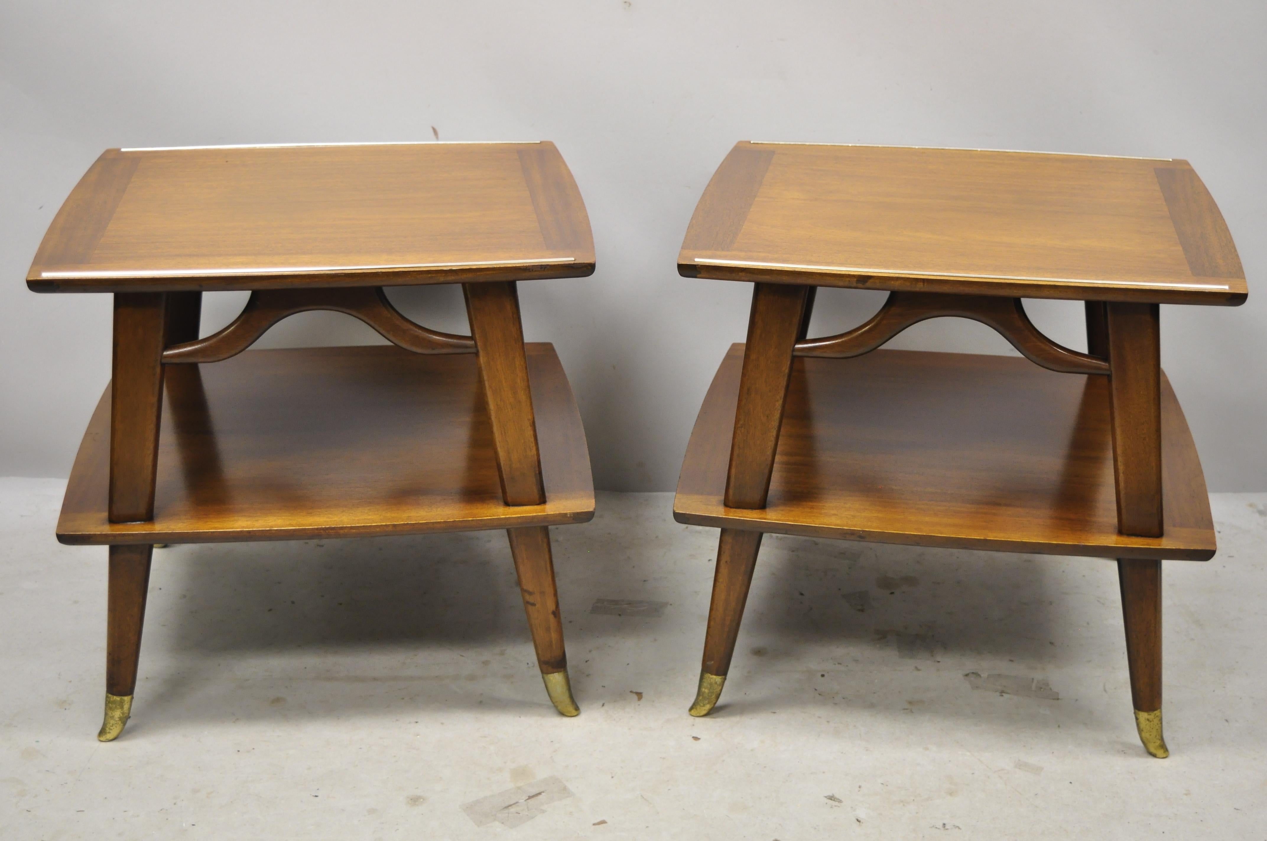 Pair of vintage Mid-Century Modern brass feet sculpted walnut 2-tier side end tables. Item features metal capped brass plated feet, lower shelf/tier, beautiful wood grain, nicely carved details, tapered legs, very nice vintage pair, sleek sculptural
