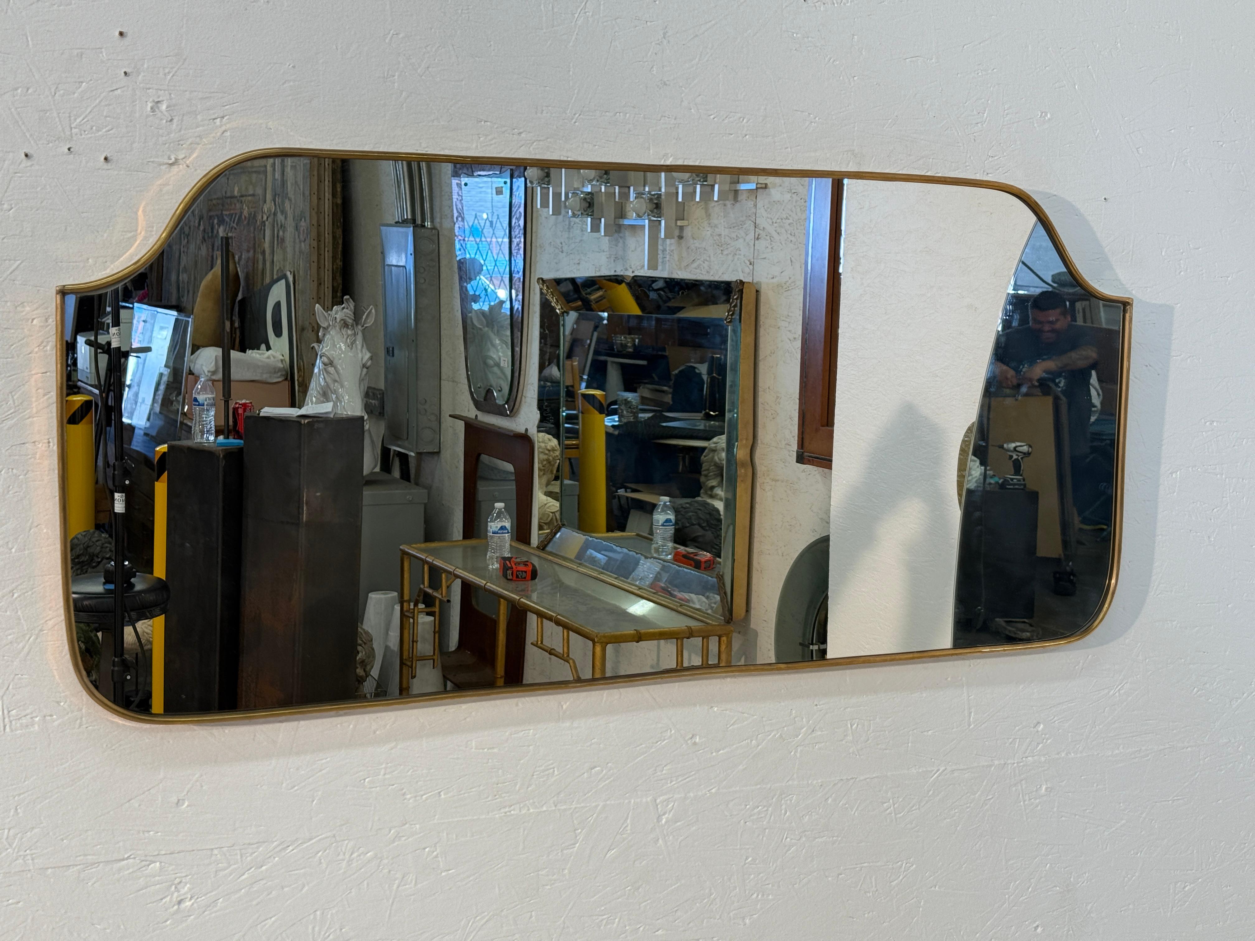 Specchio da parete vintage della metà del secolo scorso, caratterizzato da un'esclusiva forma rettangolare asimmetrica con bordi dolcemente curvi e sottili dettagli di contorno sugli angoli superiori. La cornice è realizzata in ottone con una patina