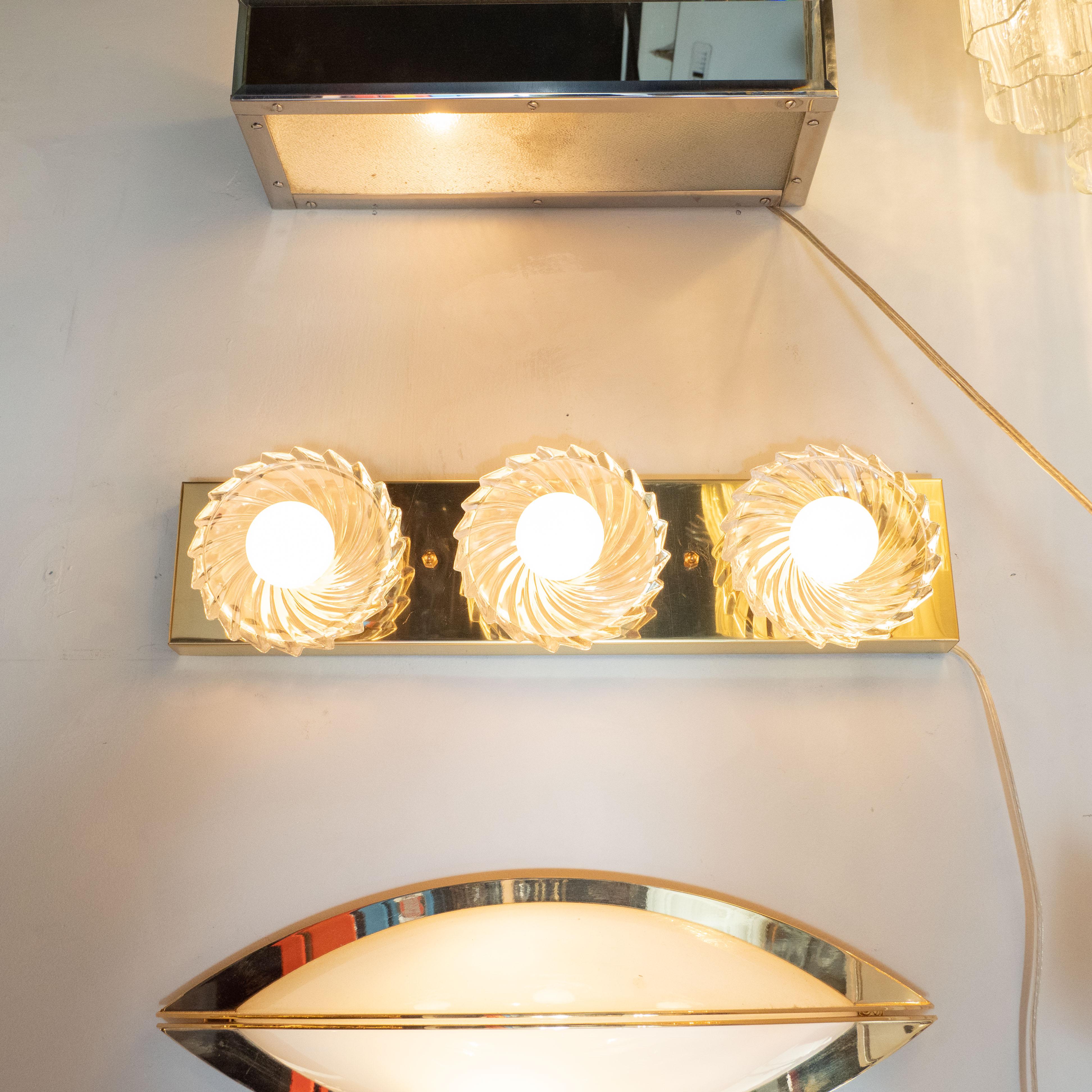 This whimsical and refined Mid-Century Modern three bulb vanity light was realized in the United States by the esteemed maker Lightolier, circa 1970. It features a volumetric polished brass plate with three circular cupped shades- resembling