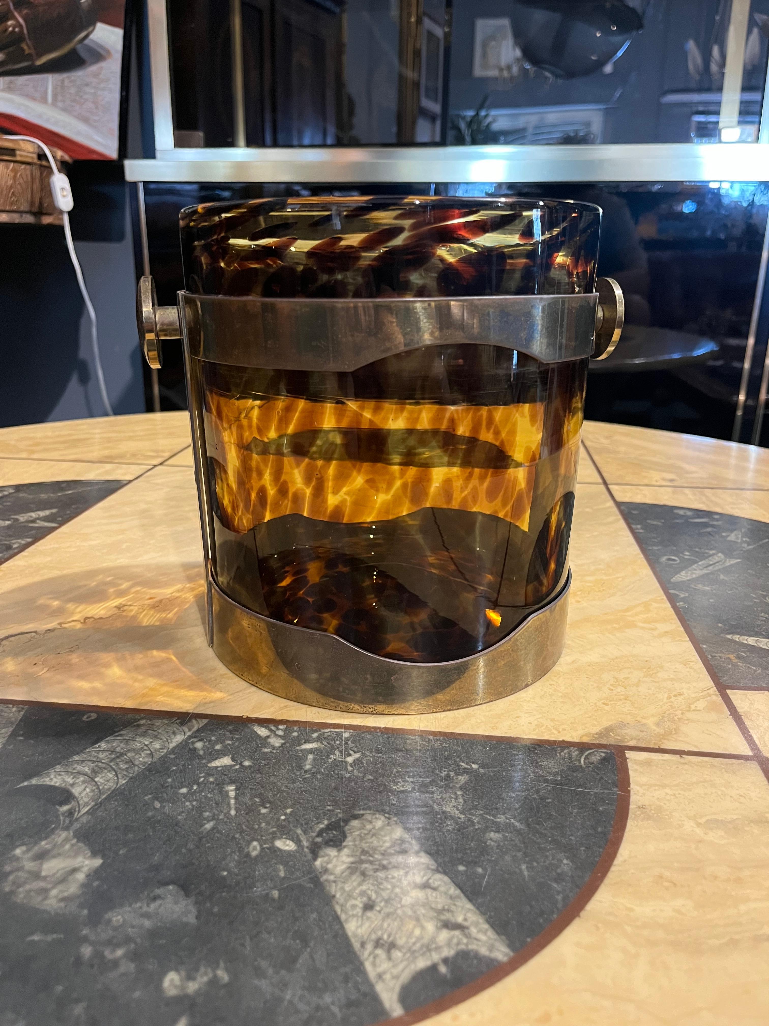This charming ice bucket is an elegant example of Guzzini design, created in the 1970s, the golden age of Italian design. The object is immediately striking for the contrast between the solid, shiny brass structure and the faux tortoiseshell-effect
