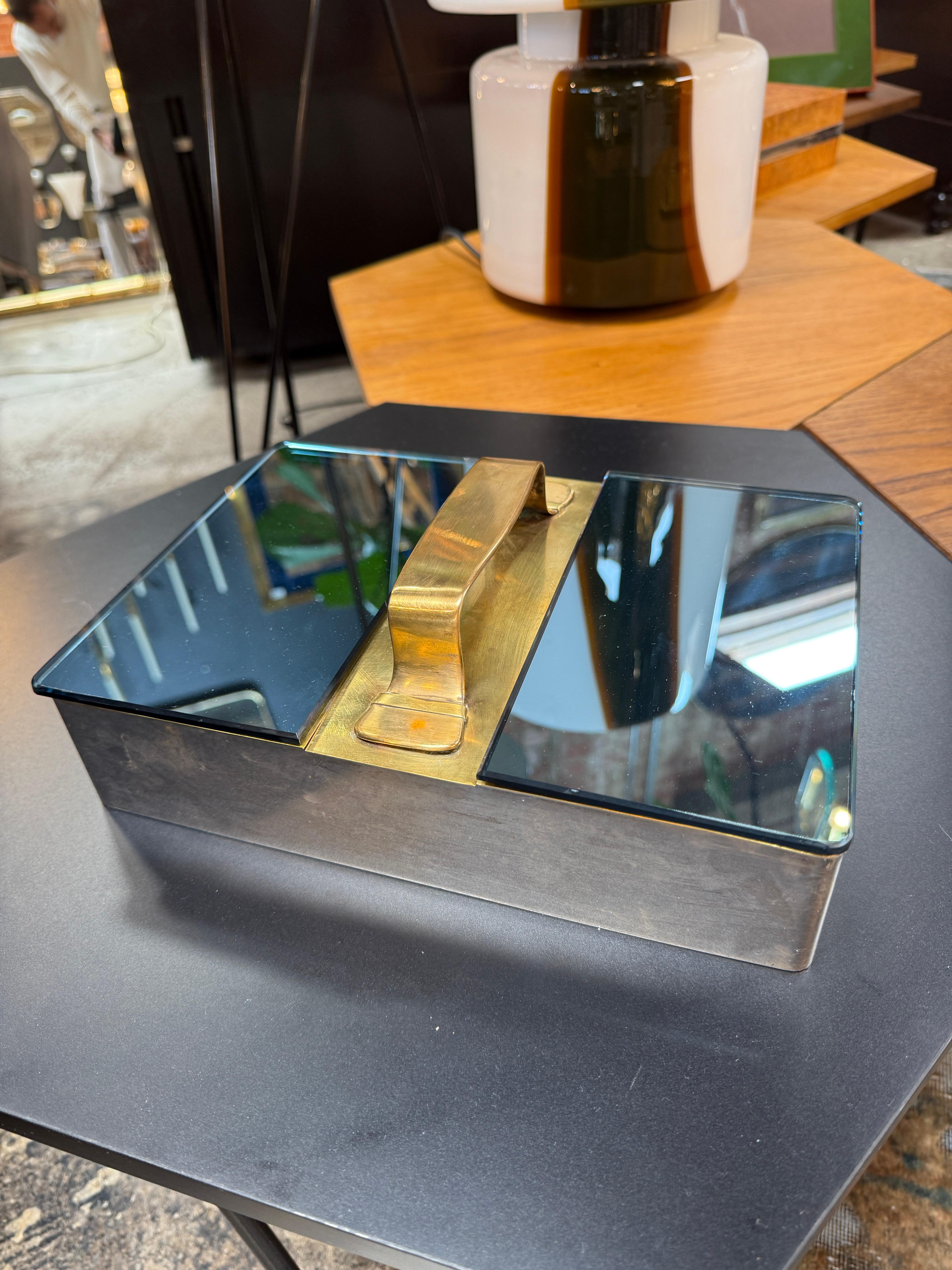 This elegant vanity/storage box showcases a striking blend of materials and craftsmanship. Featuring a polished brass handle at the center, the box is topped with beveled mirrored lids that swing open to reveal a warm, wood-lined interior. The dual