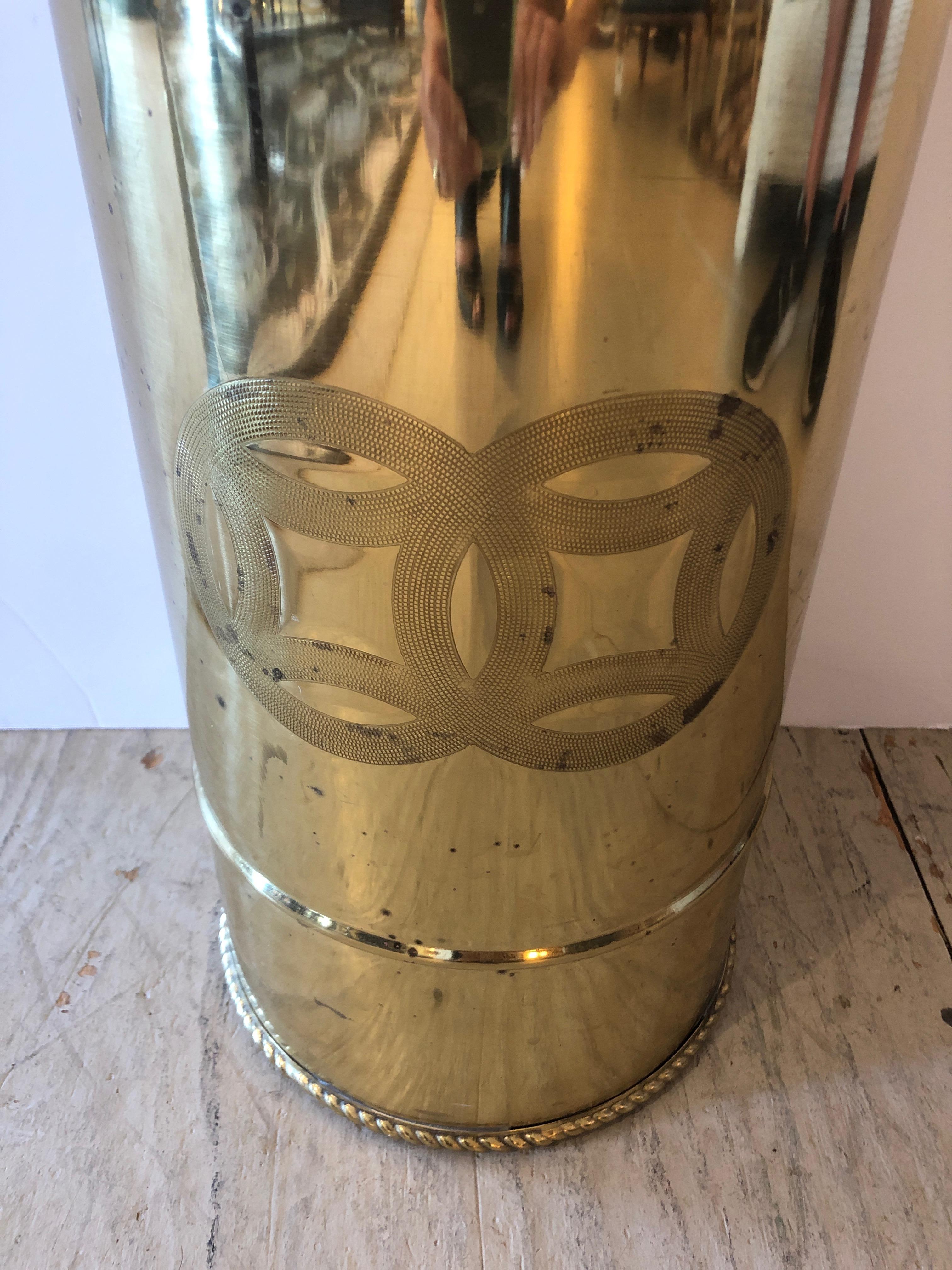 Elegant brass cylindrical umbrella stand or cane holder having decorative twisted rope at the top and bottom with central engraved geometric emblem.