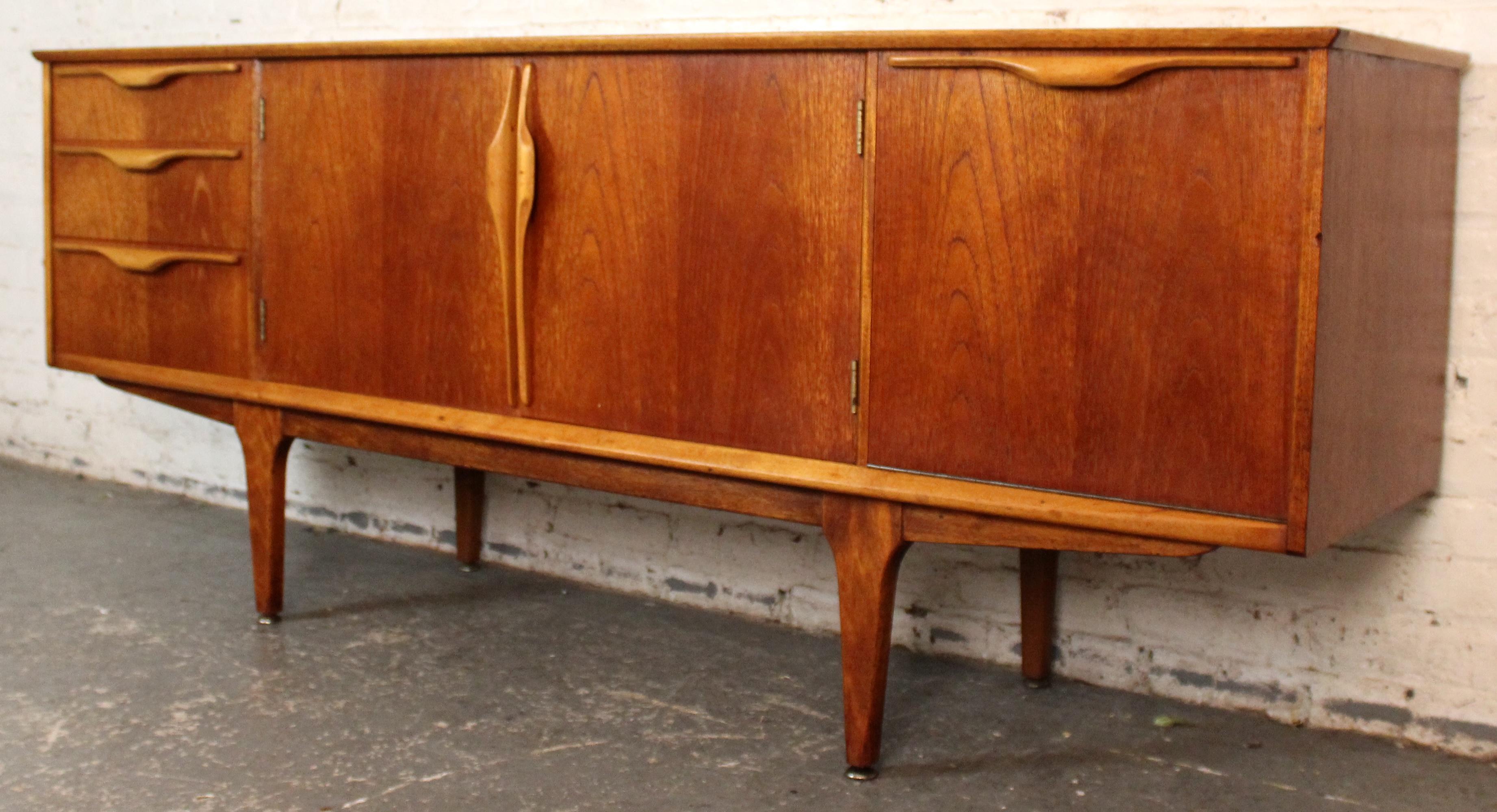 Elegante e funzionale, questa credenza Mid-Century Modern del rinomato produttore britannico Jentique presenta una combinazione splendidamente bilanciata di caldo teak e accenti contrastanti color Oak. Con le sue linee pulite, le maniglie scolpite e