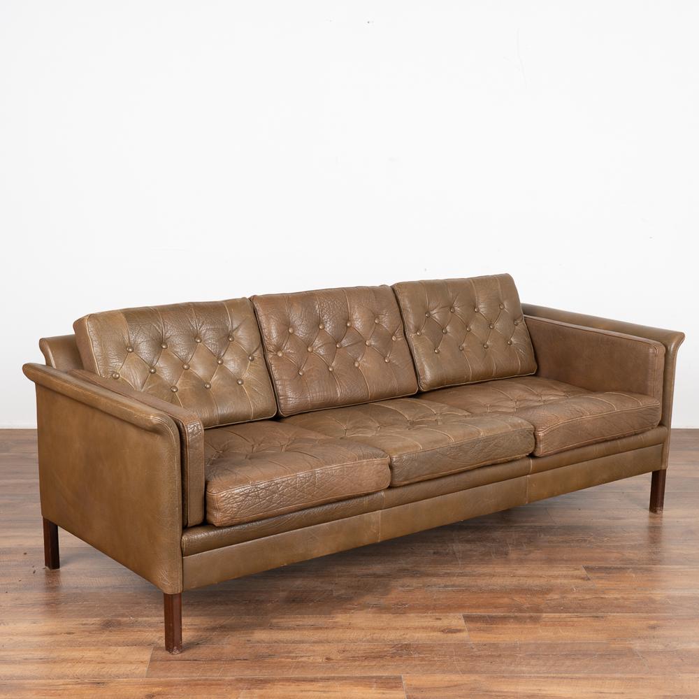 Mid-century modern brown leather three-seat sofa with button tufted back and seat cushions.
The years of use are revealed in the aged patina of the leather, including impressions, scuffs/scratches, minor discolorations, etc. which all add to the