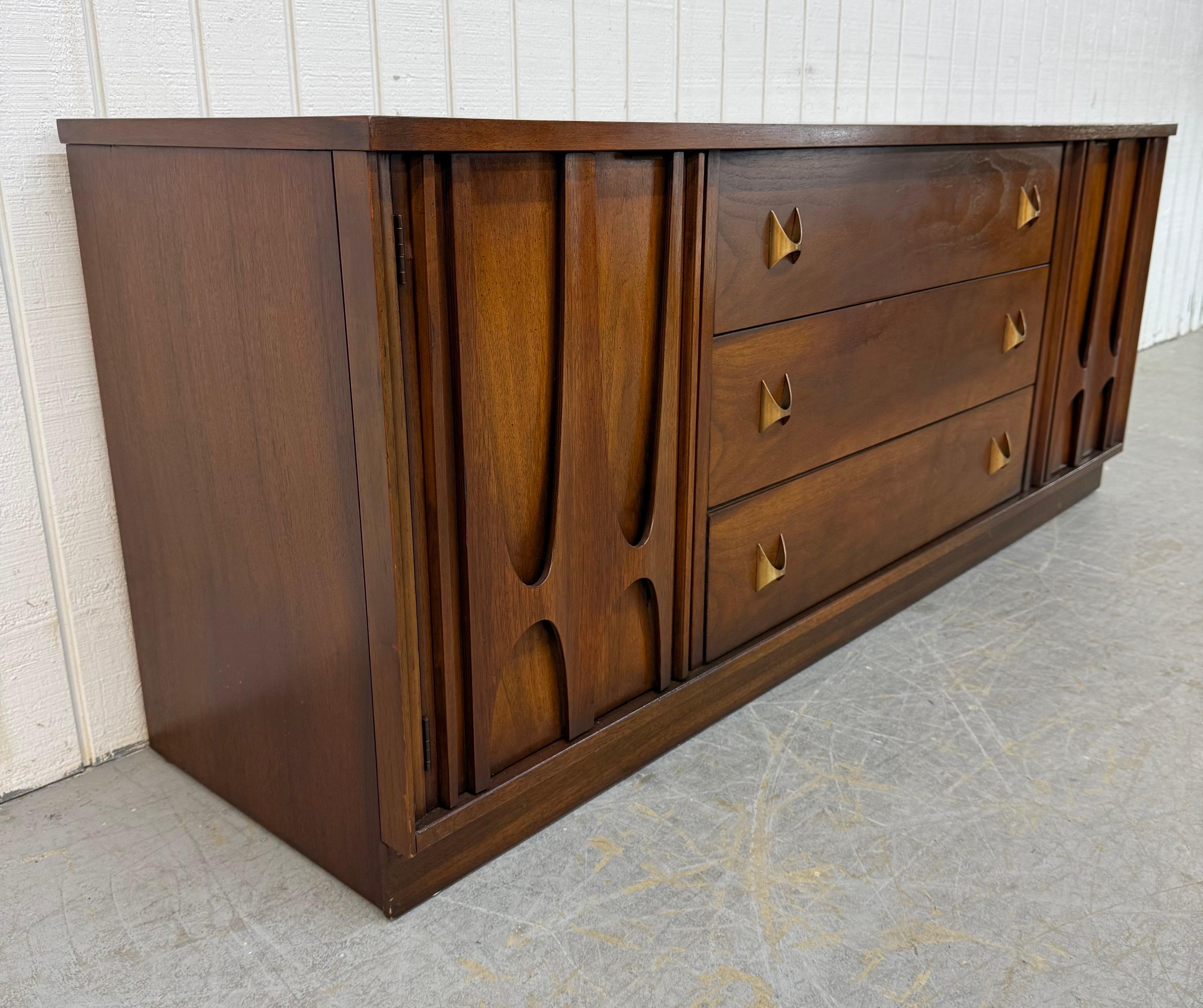 This listing is for a Mid-Century Modern Broyhill Brasilia Sideboard. Featuring a straight line design, two doors with sculpted Brasilia pulls that open up to storage space, three center drawers with original brass Brasilia hardware, plinth base,