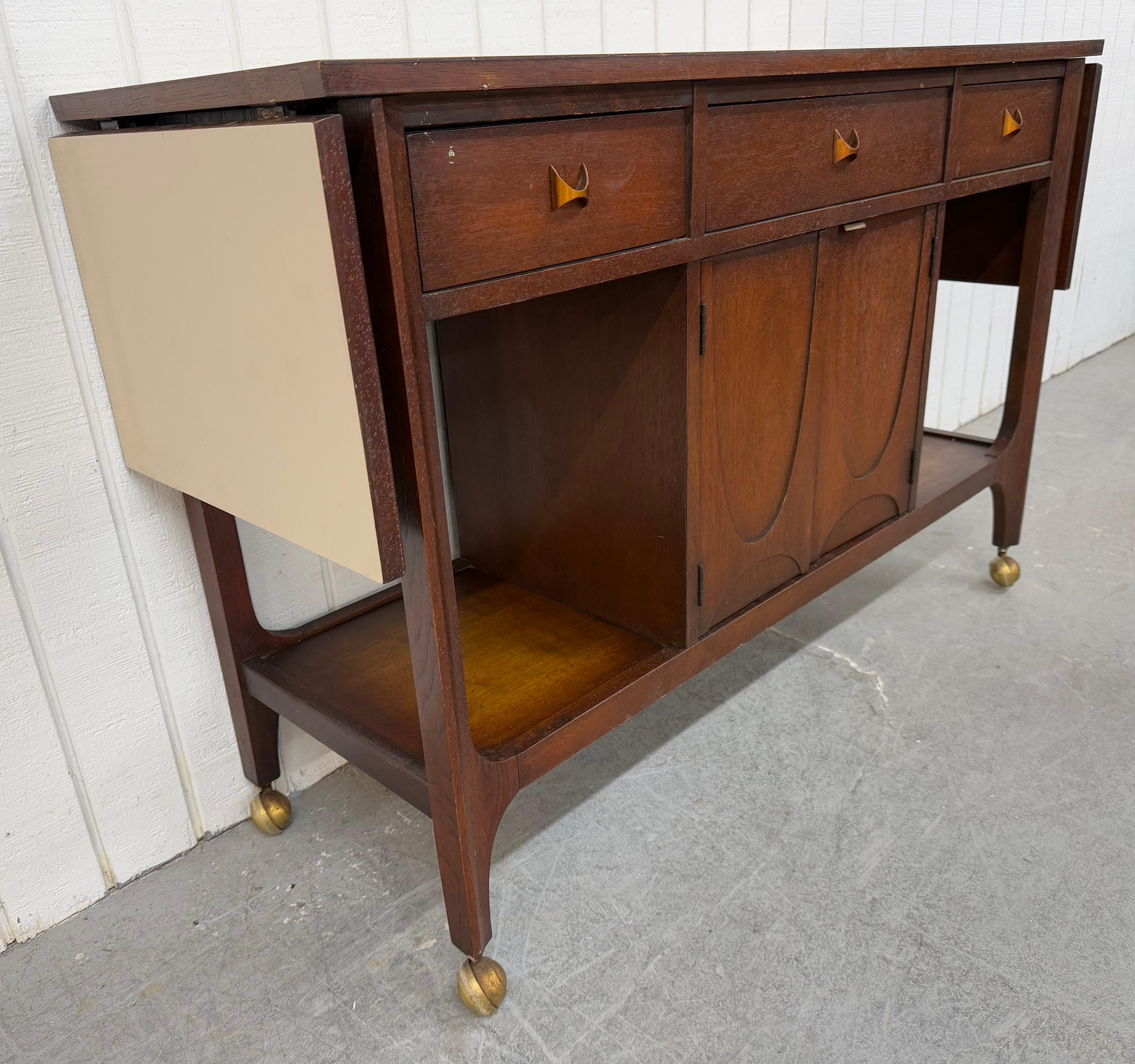 This listing is for a Mid-Century Modern Broyhill Brasilia Walnut Bar Cart. Featuring a straight line design, laminated top that expands to 70” L, three drawers with original brass Brasilia hardware, two doors that open up to storage space, modern