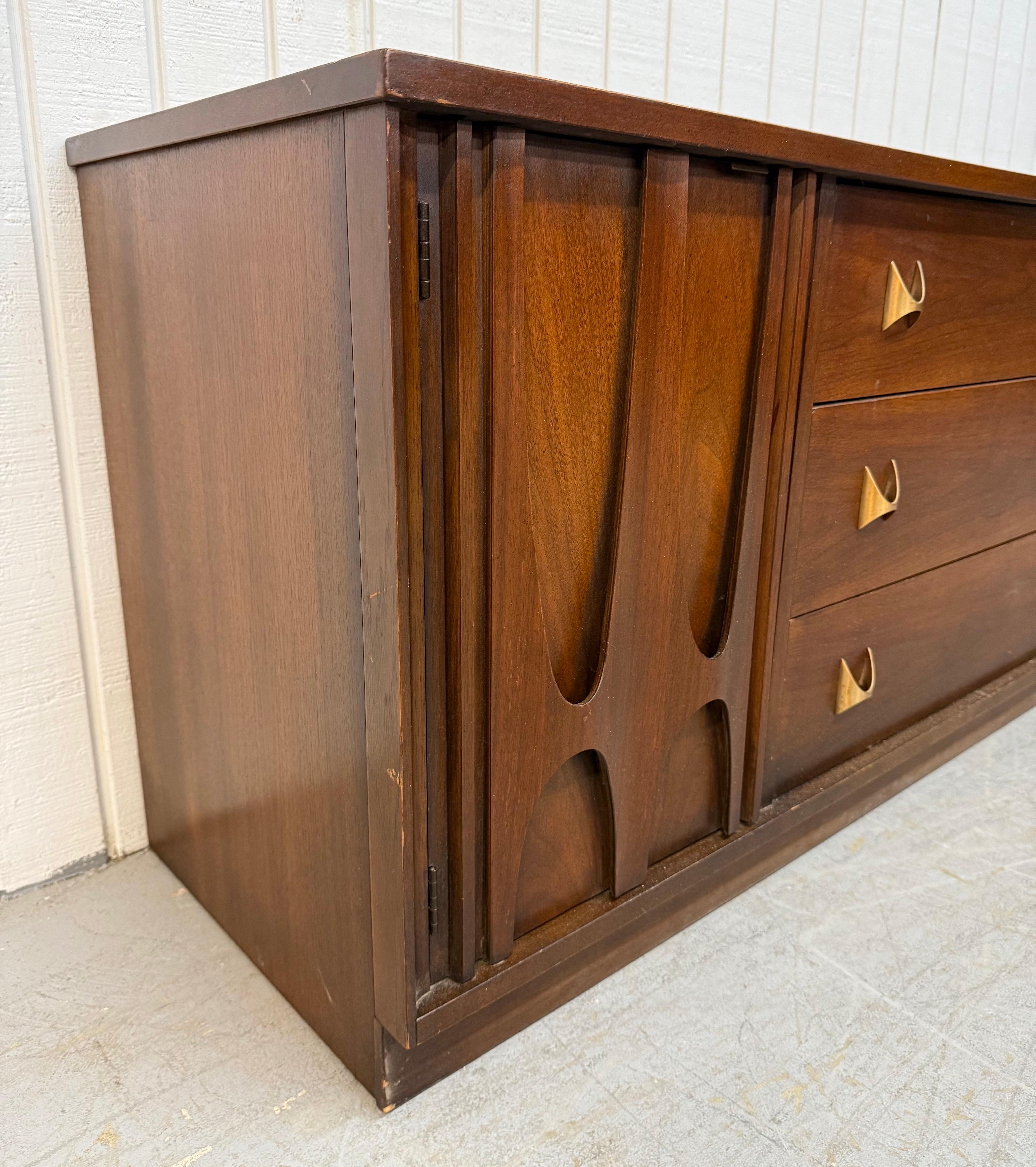 This listing is for a Mid-Century Modern Broyhill Brasilia Walnut Sideboard. Featuring a straight line design, two doors with iconic sculpted Brasilia pulls, three center drawers with the original Brasilia brass hardware, plinth base, and a